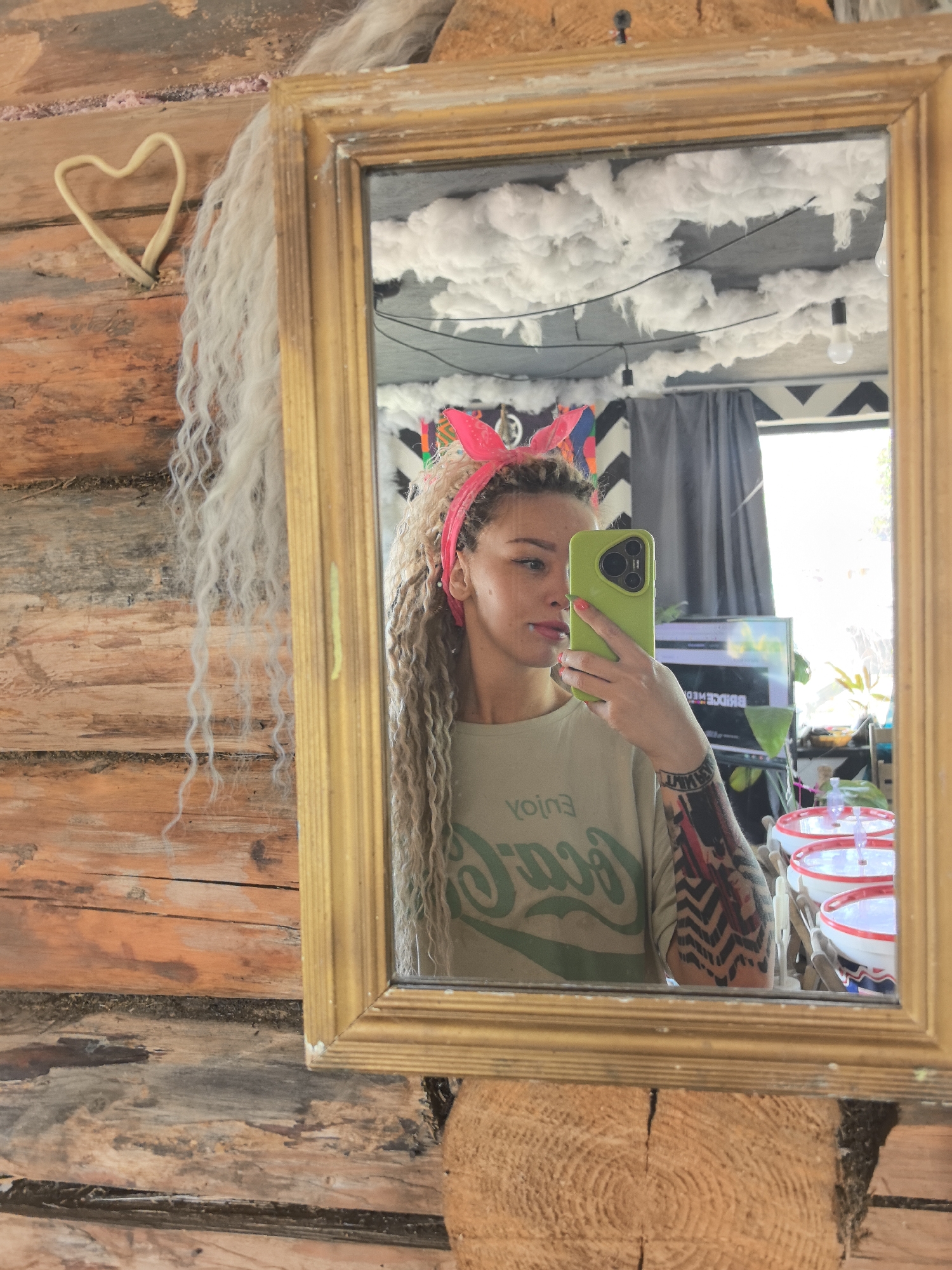 A woman is taking a mirror selfie. She is wearing a light green t-shirt with the words "enjoy Coca-Cola" printed backward on it and a pink headband with rabbit ears. She has long blonde hair and tattoos on her left arm. The mirror is framed in gold and is hanging on a wall made of logs. To the left of the mirror, there is a heart-shaped decoration made of a light-colored material. In the reflection, one can see a room with a television, plants, and a black-and-white zigzag pattern on the wall. The ceiling is decorated with fluffy white material resembling clouds with hanging lightbulbs.
The location in the city of Noviy, Russia, cannot be determined from the image.