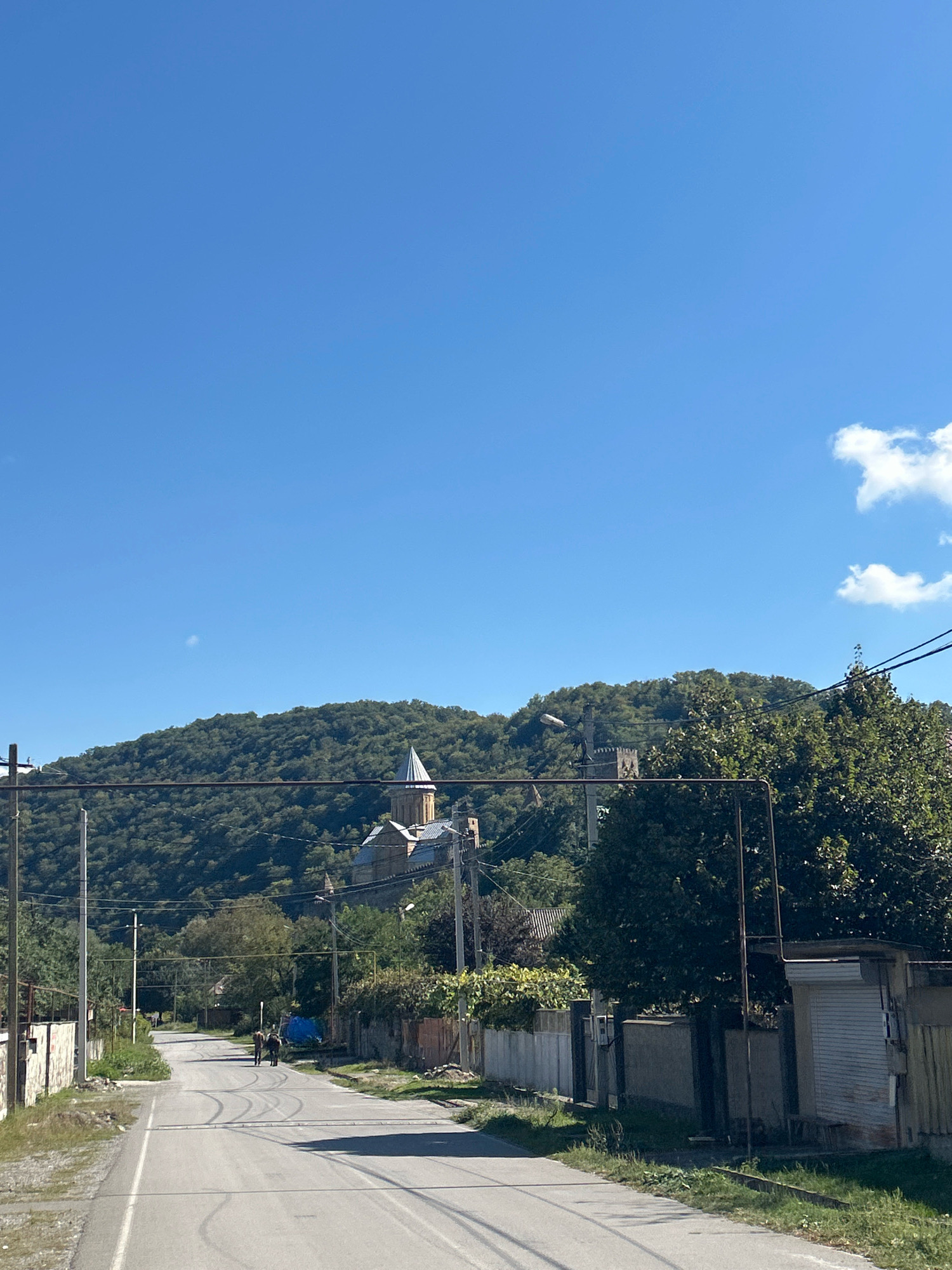 The image captures a street scene in Shalikiantkari, Georgia. A paved road stretches forward, flanked by fences and vegetation on both sides. Two figures, appearing to be people, are walking away from the camera on the road. In the background, a lush, tree-covered hill rises, with a prominent building, likely a church or monastery, situated on its slopes. The sky above is a clear blue with a few wispy clouds. The overall impression is a rural or semi-rural setting bathed in daylight.