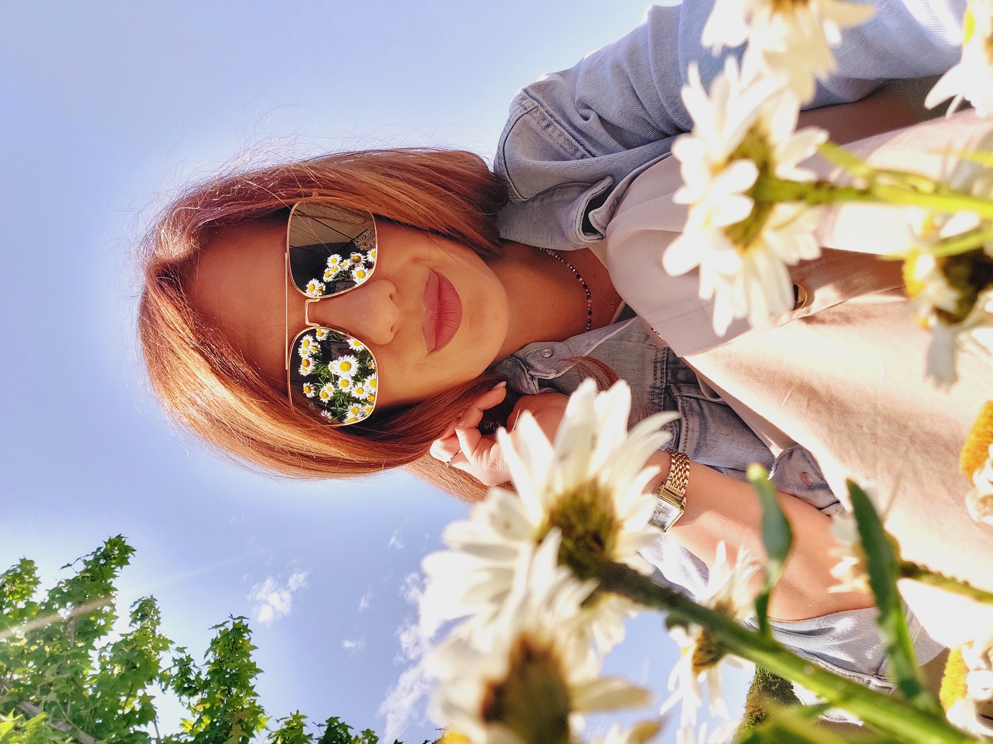 A low-angle, medium shot captures a woman with auburn hair and sunglasses, reflecting a field of daisies, as she sits outdoors on a bright, sunny day. She is wearing a light blue denim jacket over a cream-colored top and beige pants. A gold watch adorns her left wrist. Daisies are in the foreground, out of focus, obscuring the lower portion of the frame. The background shows a clear blue sky with some clouds and the green leaves of a tree. The scene appears to be in a park or garden, possibly in Sofia, Bulgaria, given the sunlight and clear sky, but no specific landmarks are visible.