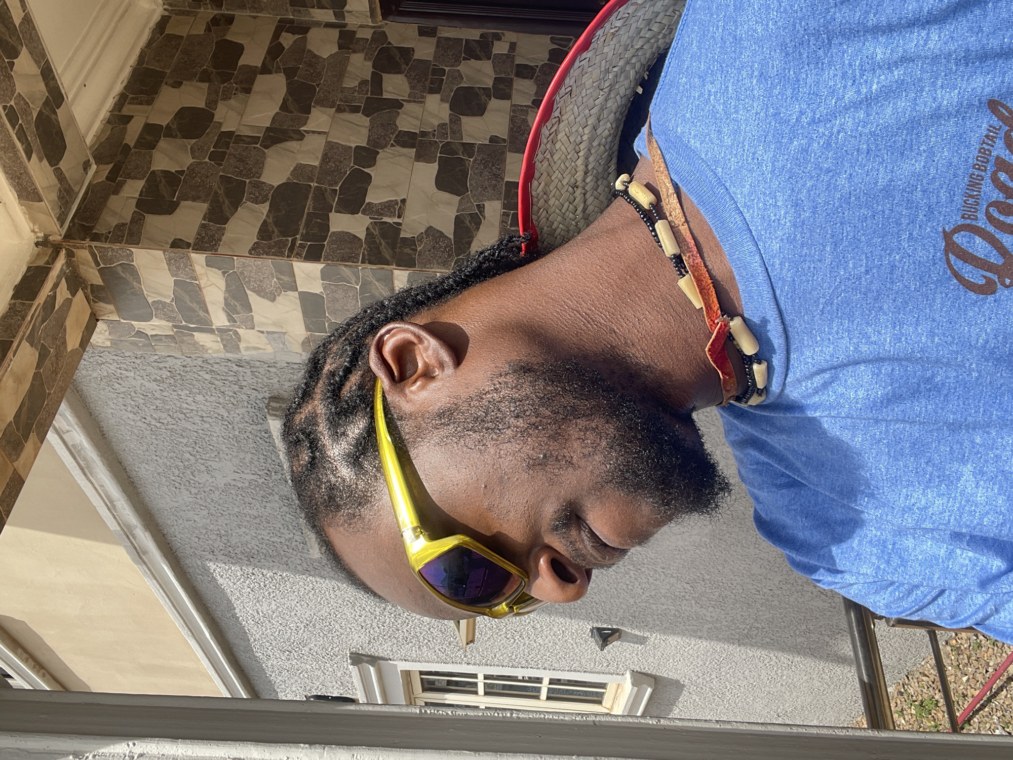 A medium-shot image shows a bearded Black man with cornrow braids and gold sunglasses, wearing a blue t-shirt and a necklace with bone and black beads. He is outdoors, likely in front of a building with tiled walls and a small window. The text on the t-shirt reads "BUCKING BOBTAIL." Given the tiled facade and general appearance, the location is possibly in a residential area of Abuja, Nigeria.