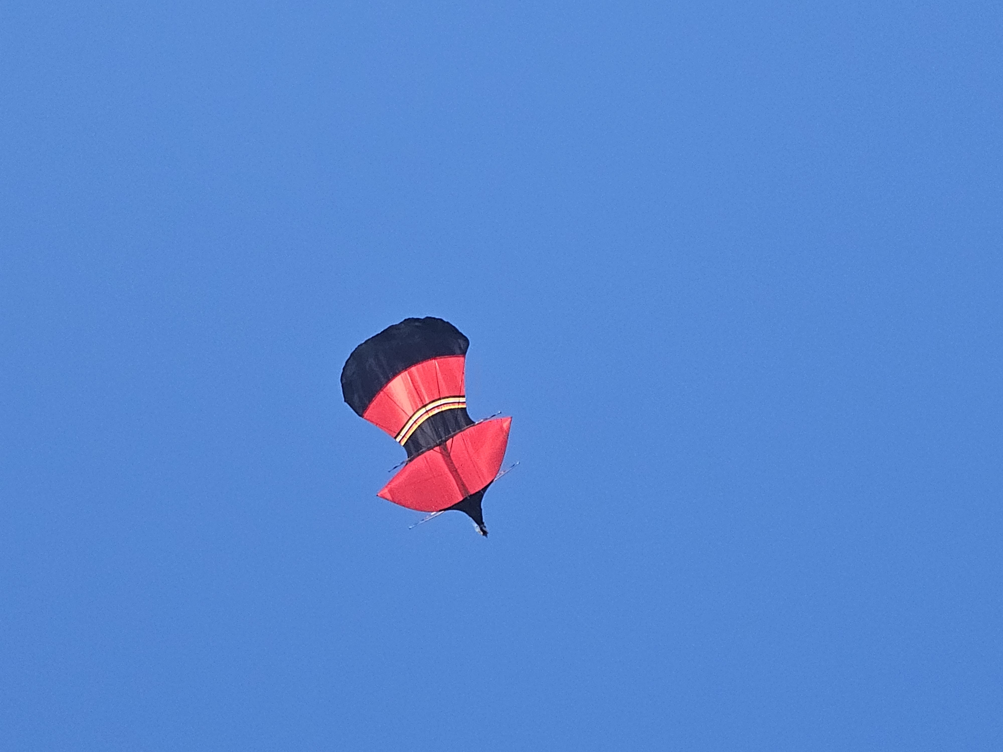 A red and black kite is flying in a clear, blue sky. The kite is shaped like a top, with a round, black top, a section with red and yellow stripes, and a red bottom. The kite is located in Denpasar, Indonesia.