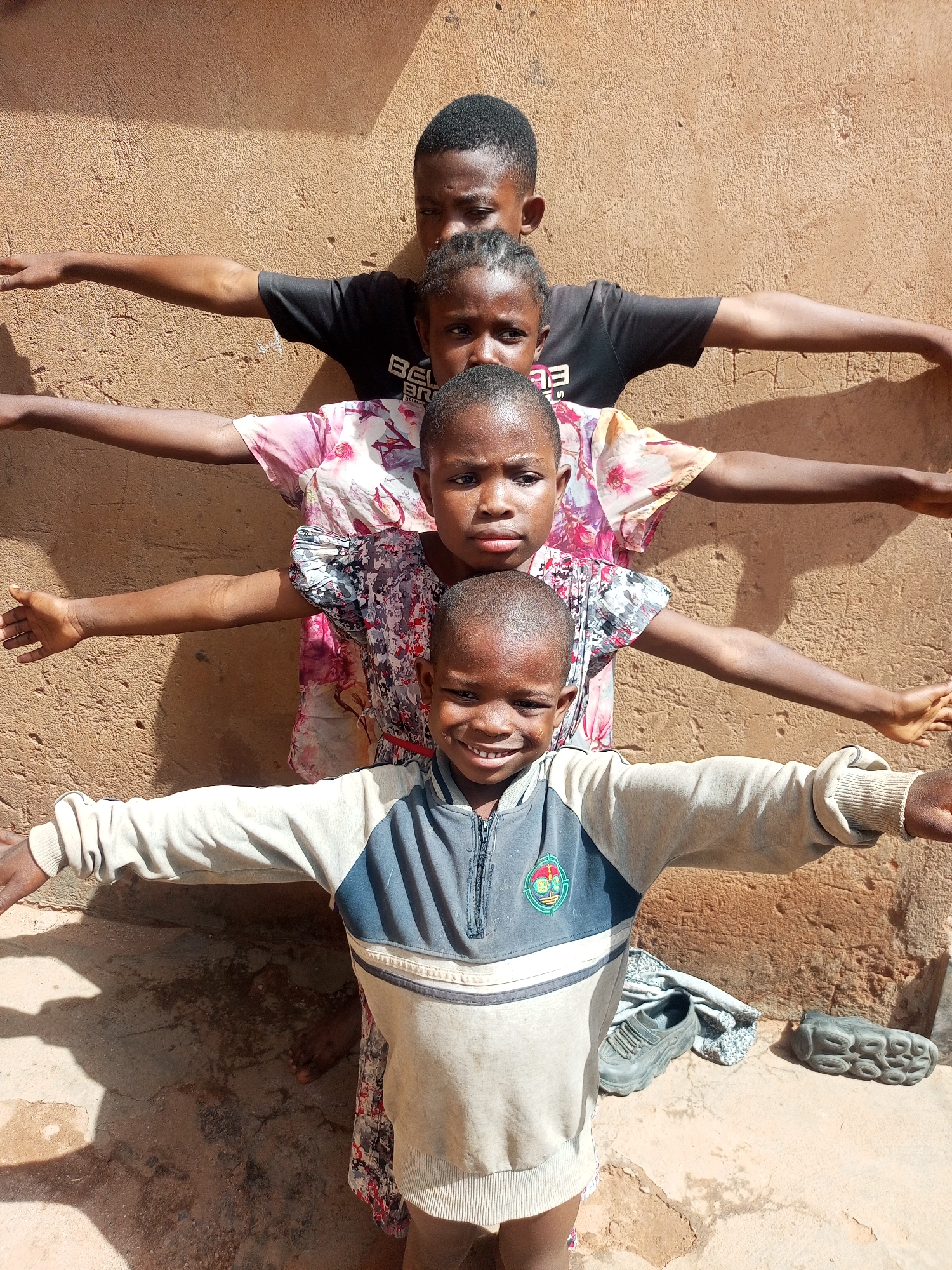 Four children of African descent stand in a row against a textured, adobe-colored wall in an outdoor setting that may be Tempure, Nigeria, given the prompt. Each child has their arms extended to the sides in a horizontal line. From top to bottom, the children are arranged as follows: a boy wearing a black t-shirt, a girl with braided hair, a girl in a floral dress, and a younger boy wearing a long-sleeved shirt with blue and white panels. The young boy at the bottom is smiling. Shadows are cast on the ground in front of the children, where a shoe and a sandal lie. The ground appears to be concrete with cracks and dirt.