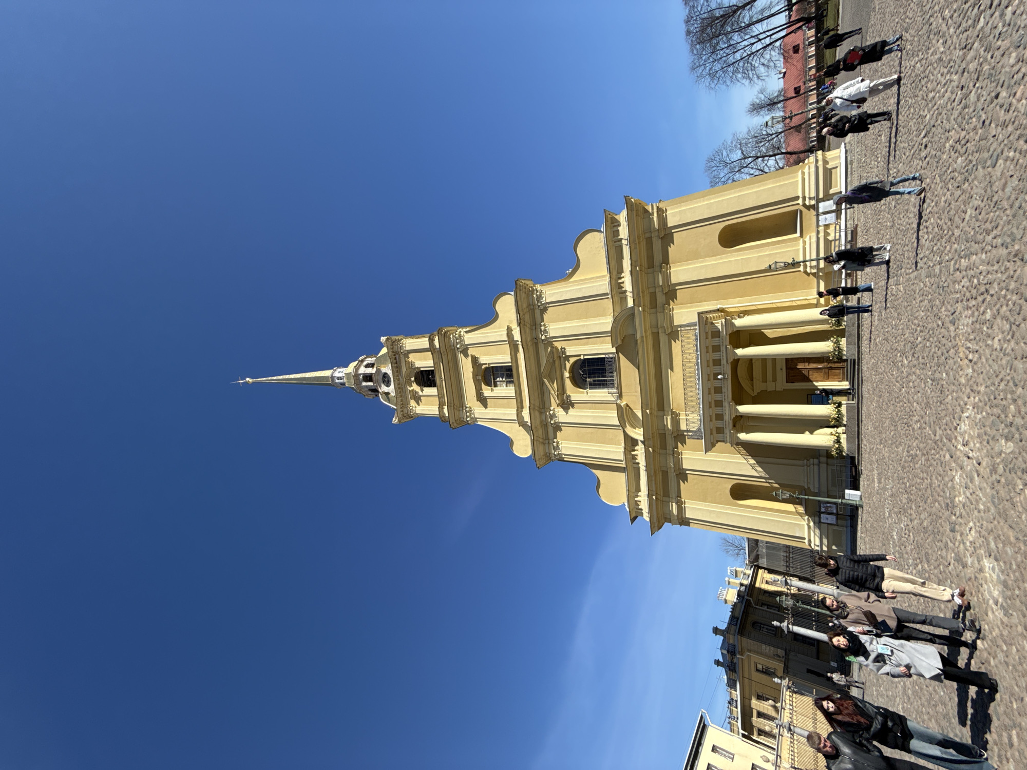 The image shows the Peter and Paul Cathedral in Saint Petersburg, Russia. The cathedral, a tall, yellow building, is positioned vertically in the frame against a clear, blue sky. Several people are standing on a cobblestone surface in front of the cathedral. A row of buildings is visible behind the people, and bare trees are visible near a red building in the upper right corner.