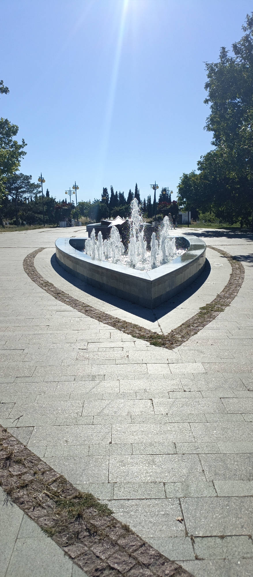 An outdoor shot shows a heart-shaped fountain with water spraying upwards in the foreground. The fountain is surrounded by a paved area with a pattern of stones and grass. In the background, there are trees, streetlights, and a clear blue sky. The sun is shining brightly, with a visible lens flare.