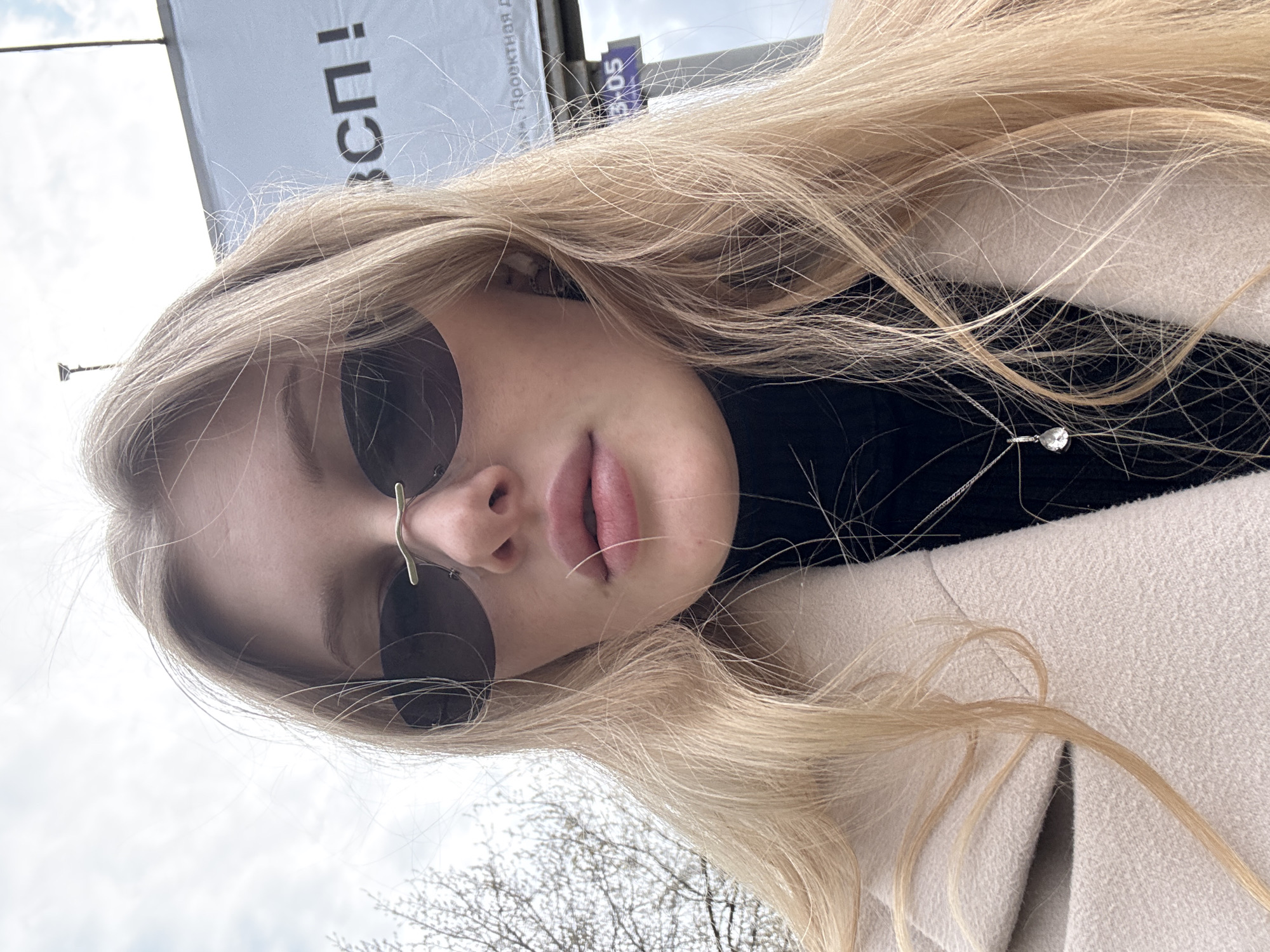 A selfie photograph depicts a woman with blonde hair and sunglasses, wearing a black turtleneck sweater and a light-colored coat. A thin silver necklace with a pendant is visible around her neck. In the background, part of a billboard with Russian text is visible. Based on the text on the billboard (Проектная д), this image may have been taken in Perm, Russia. The sky is overcast, and bare tree branches are visible in the bottom part of the image. The woman appears to be standing outside.
