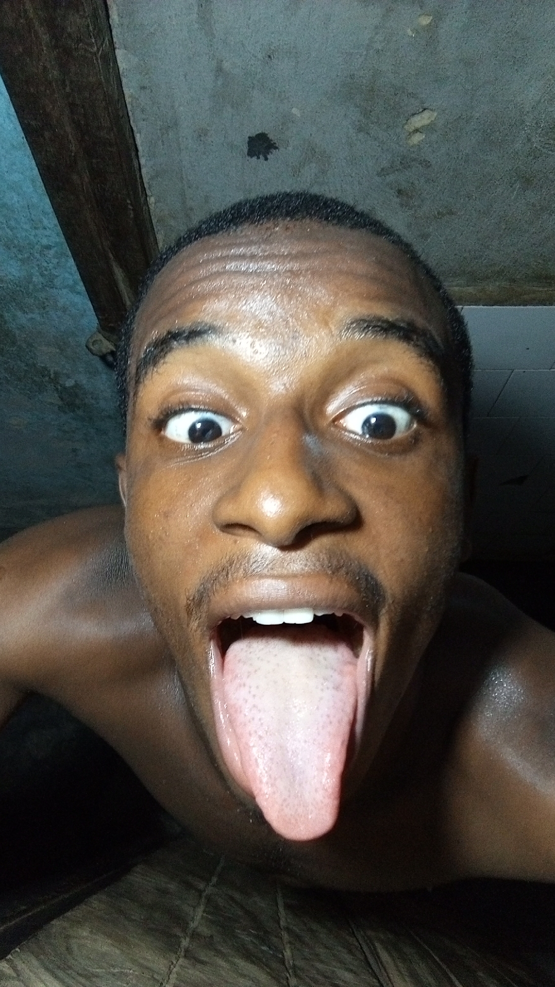 A close-up, low-angle shot features a person, likely male, with dark skin, making an exaggerated, playful face. His head is cropped just above the forehead, revealing short, dark hair and a thin mustache. His eyes are wide open, pupils dilated, and eyebrows are raised, creating an expression of surprise or intensity. His mouth is open wide, with his tongue fully extended downwards, showing its textured surface. The person's skin appears somewhat shiny, reflecting the dim light.
The background above the person's head consists of a textured, light grey or blue-toned wall or ceiling, featuring some dark stains or marks. A dark, thick wooden beam or structural element runs diagonally across the upper left portion of the frame. The area below the person's chin and tongue is dark, with a subtly textured surface, possibly a floor or another object. The lighting in the scene is dim and uneven, suggesting an indoor setting with low or artificial light. No text is visible. The overall mood of the image is informal and humorous due to the exaggerated facial expression.