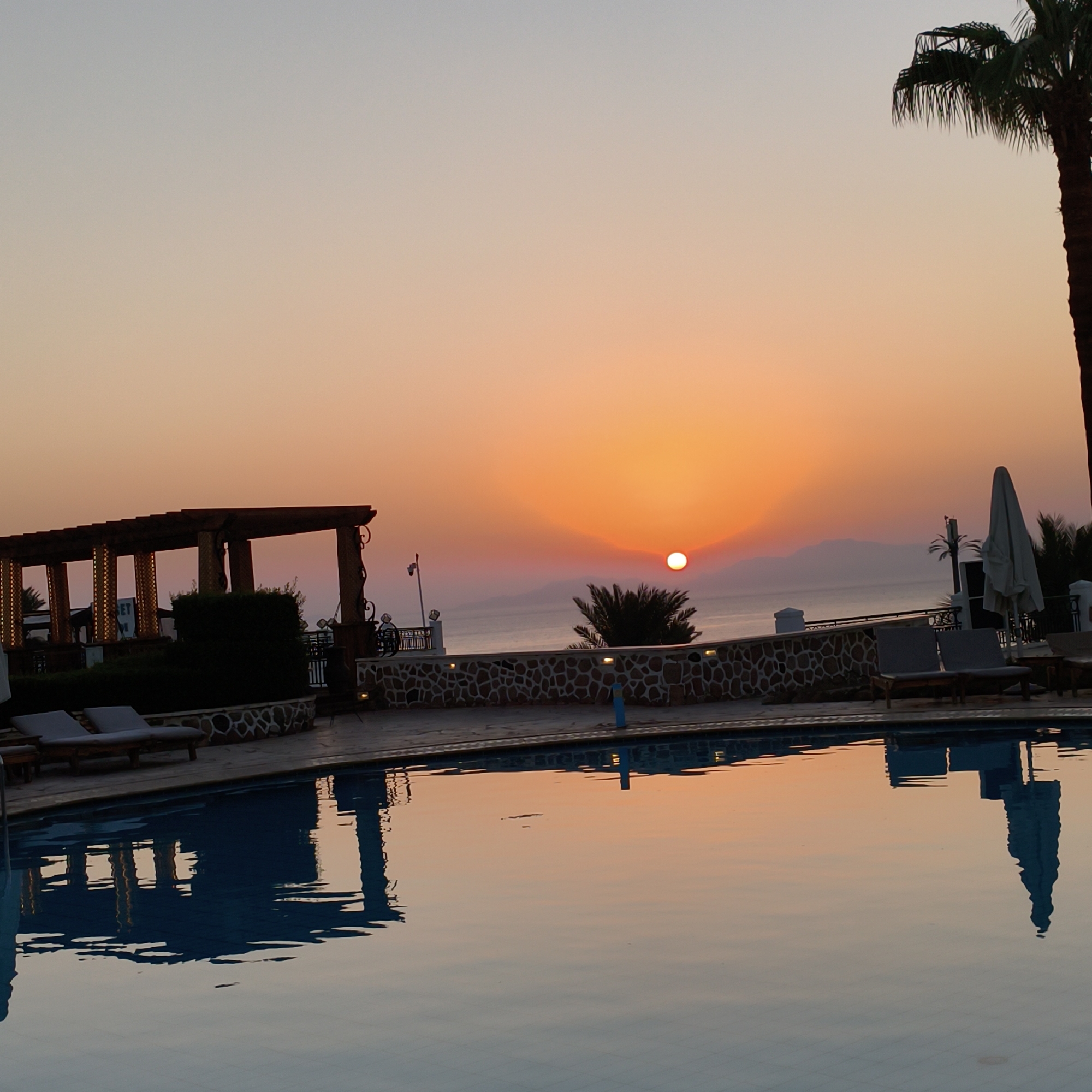 The image depicts a serene sunset scene at a resort, featuring a swimming pool in the foreground and the ocean in the background. The sun is setting over the ocean, casting a warm orange glow across the sky and reflecting on the water's surface. Palm trees are visible on the right side of the image, adding to the tropical ambiance. The swimming pool is empty, with lounge chairs set up by its side, suggesting a peaceful and relaxing atmosphere. A pergola with decorative columns is situated near the pool, offering shade and architectural interest. The scene is characterized by the soft light of dusk, with the water of the pool mirroring the colors of the sky and the surrounding structures.