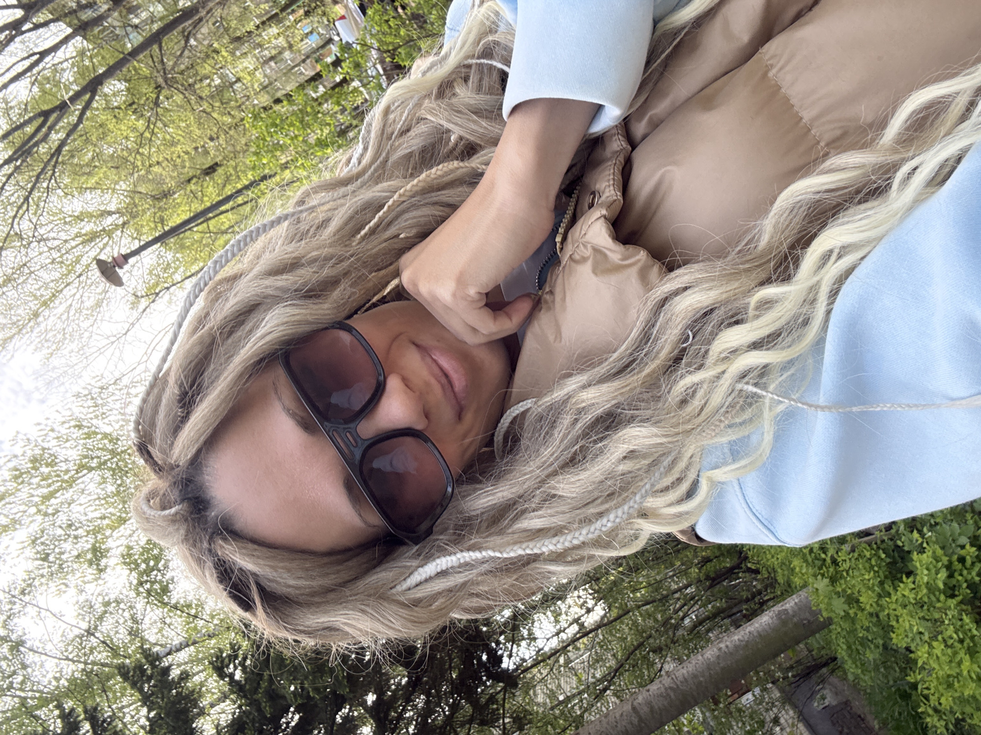 A woman with blonde braided hair, wearing sunglasses, a light blue hoodie, and a tan puffer vest, is posing for a selfie outdoors. She is holding her hand near her face. Trees and a street lamp are visible in the background, suggesting an urban park setting. The location is in the city of Unknown, Unknown.