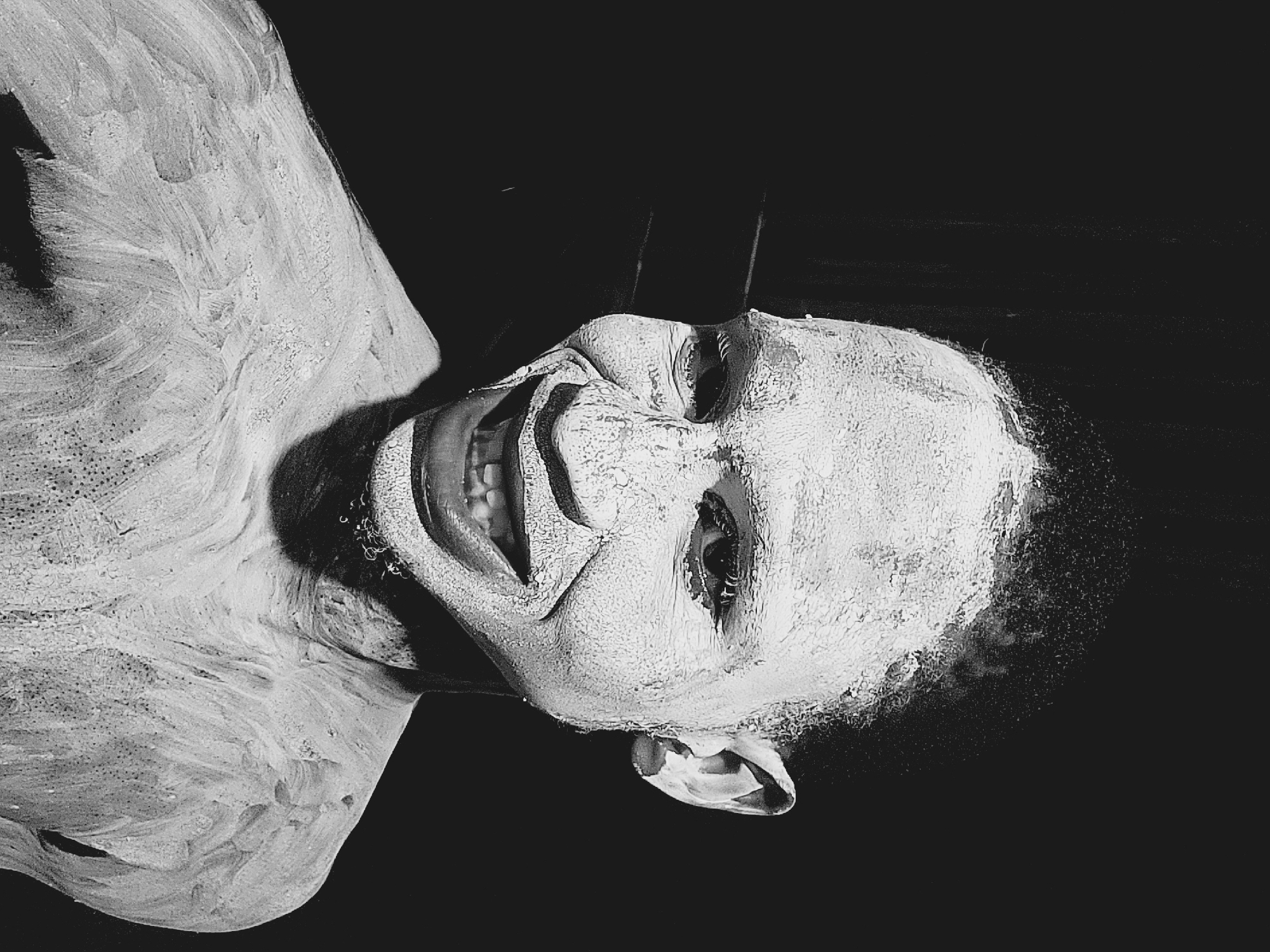 A black and white photograph captures a person, likely male, whose head and upper torso are heavily covered in a thick, textured white substance, resembling paint or plaster. The image is oriented sideways, with the person's head positioned towards the right side of the frame, suggesting a reclining or unconventional pose.
The subject displays a wide, exaggerated grin, revealing their teeth, while their eyes are open and directed away from the viewer. This theatrical facial expression is amplified by the white coating, which highlights the contours and creases of the face, creating a dramatic and somewhat unsettling effect. The white substance shows visible brushstrokes or an uneven, slightly cracked texture across the skin, hair, and visible parts of the body.
The background is a uniform, deep black, which provides a stark contrast and isolates the figure, drawing all attention to the subject. Strong, directional lighting emphasizes the textures of the white covering and casts dramatic shadows, further enhancing the intensity of the scene. The overall impression is artistic, avant-garde, and eerie, evoking performance art or a raw, expressive portrait. There is no visible text in the image.