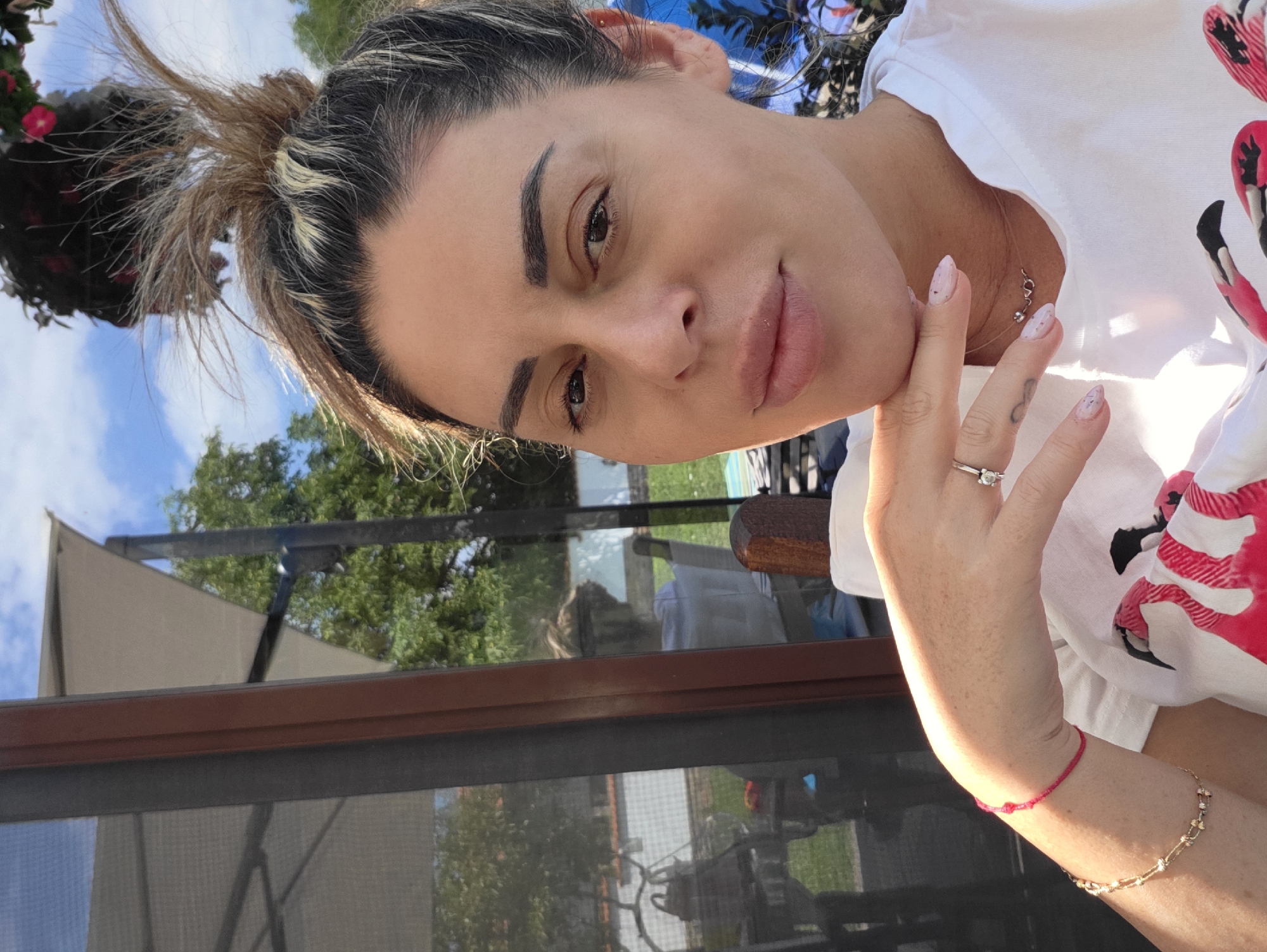 A woman is captured in a close-up, selfie-style shot outdoors in Hrabrino, Bulgaria. She is looking directly at the camera with her hand resting on her chin. She has dark hair pulled up in a messy bun and is wearing a white t-shirt with a pink flamingo graphic. Her fingernails are painted pink with white speckles, and she is wearing a ring on her left ring finger and several bracelets on her left wrist, one of which is red. A delicate necklace is visible around her neck.
The background suggests a residential outdoor setting. To the left, a glass door or window reflects an outdoor scene with trees, a patio umbrella, and furniture. In the upper right corner, a hanging flower basket is visible against a blue sky with white clouds. The overall lighting indicates it is daytime, with sunlight casting shadows and highlighting the subject.