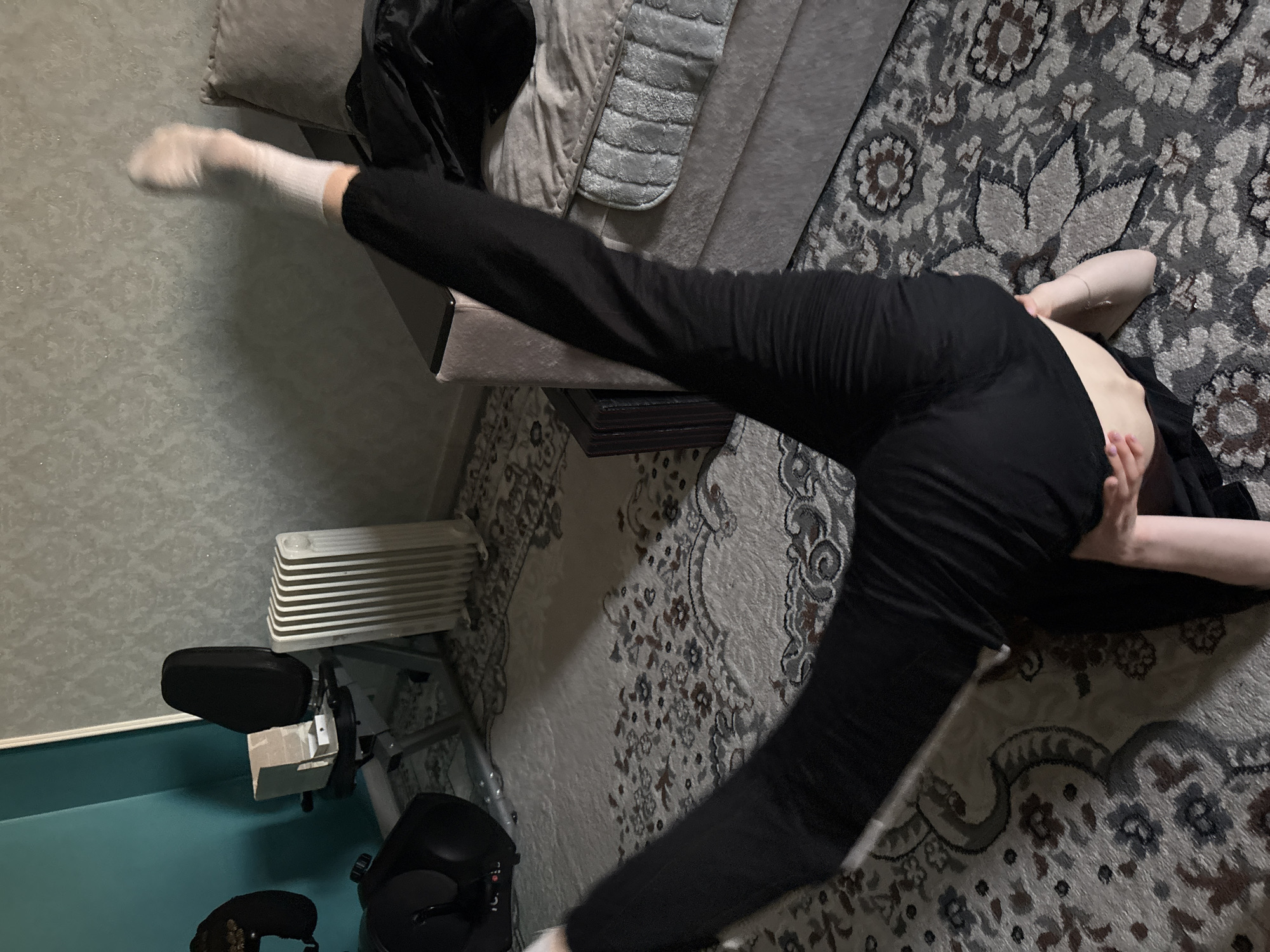 In a dimly lit room, a person is captured mid-motion, performing a high leg kick. They are dressed in black pants and white socks, and their body is arched as they extend their left leg upwards, parallel to the floor. Their right leg is bent, and their body is angled on a patterned rug.
The room appears to be a living space, with a plush sofa covered in grey blankets visible in the background. A dark table with a reflective surface is positioned near the sofa. To the left of the frame, a white electric heater with multiple fins is situated next to a wall. Further to the left, a piece of exercise equipment with black padding is partially visible.
The overall impression is one of dynamic movement and physical exertion, possibly during a dance or martial arts practice. The lighting is subdued, casting shadows and creating a sense of intimacy. The background elements suggest a home environment, though specific details about the time of day or weather are not discernible. No visible text is present in the image.
