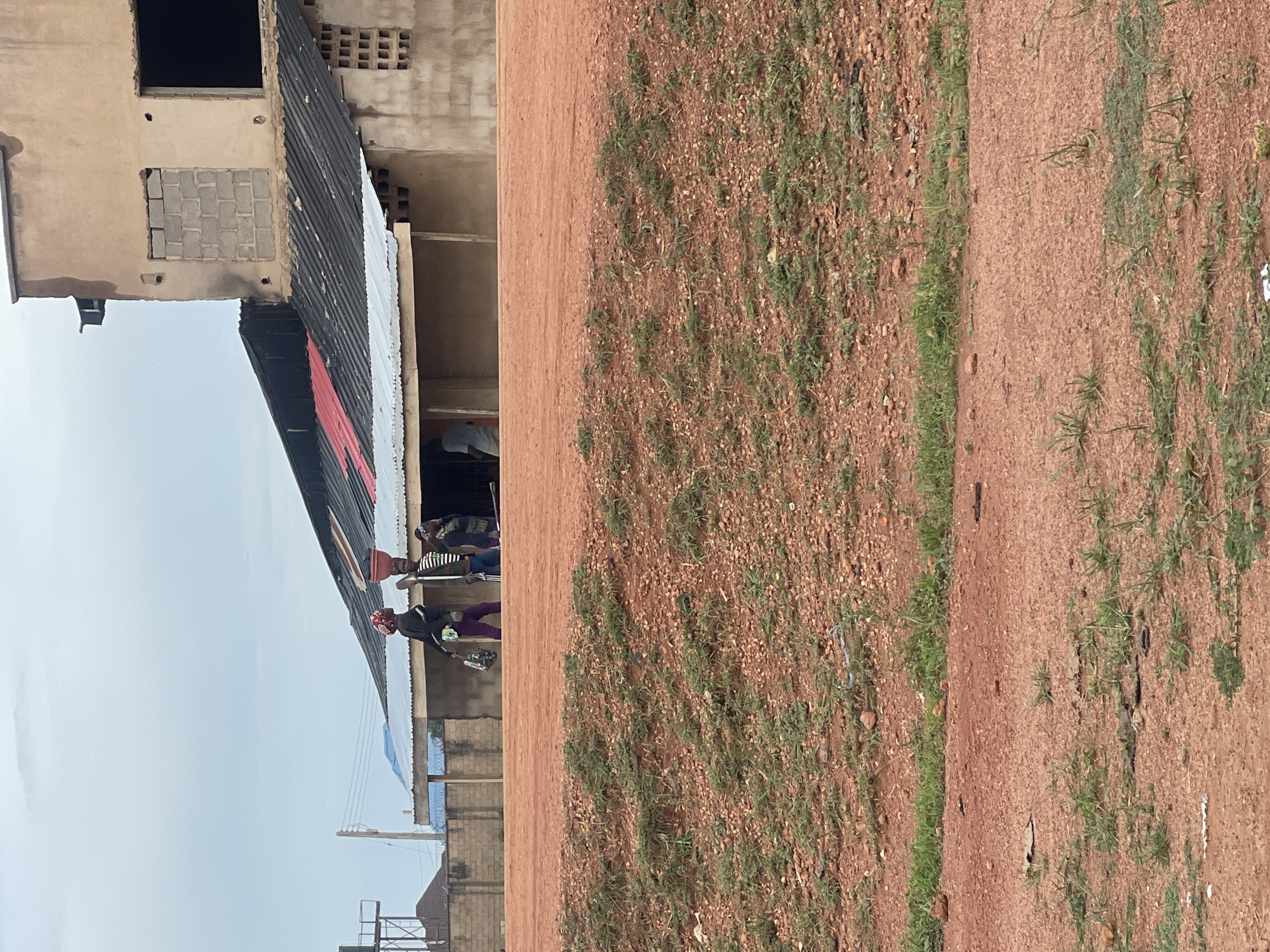 In Kwang, Nigeria, a group of people are seen near a building with a corrugated iron roof. One person is balancing a red bucket on their head, and another is carrying a shopping bag. The building appears to be under construction or in disrepair, with exposed cinder blocks and unfinished windows. The surrounding environment is dry and dusty, with sparse vegetation and reddish-brown soil. The sky is overcast, suggesting a cloudy day. The scene depicts a snapshot of daily life in a Nigerian community.