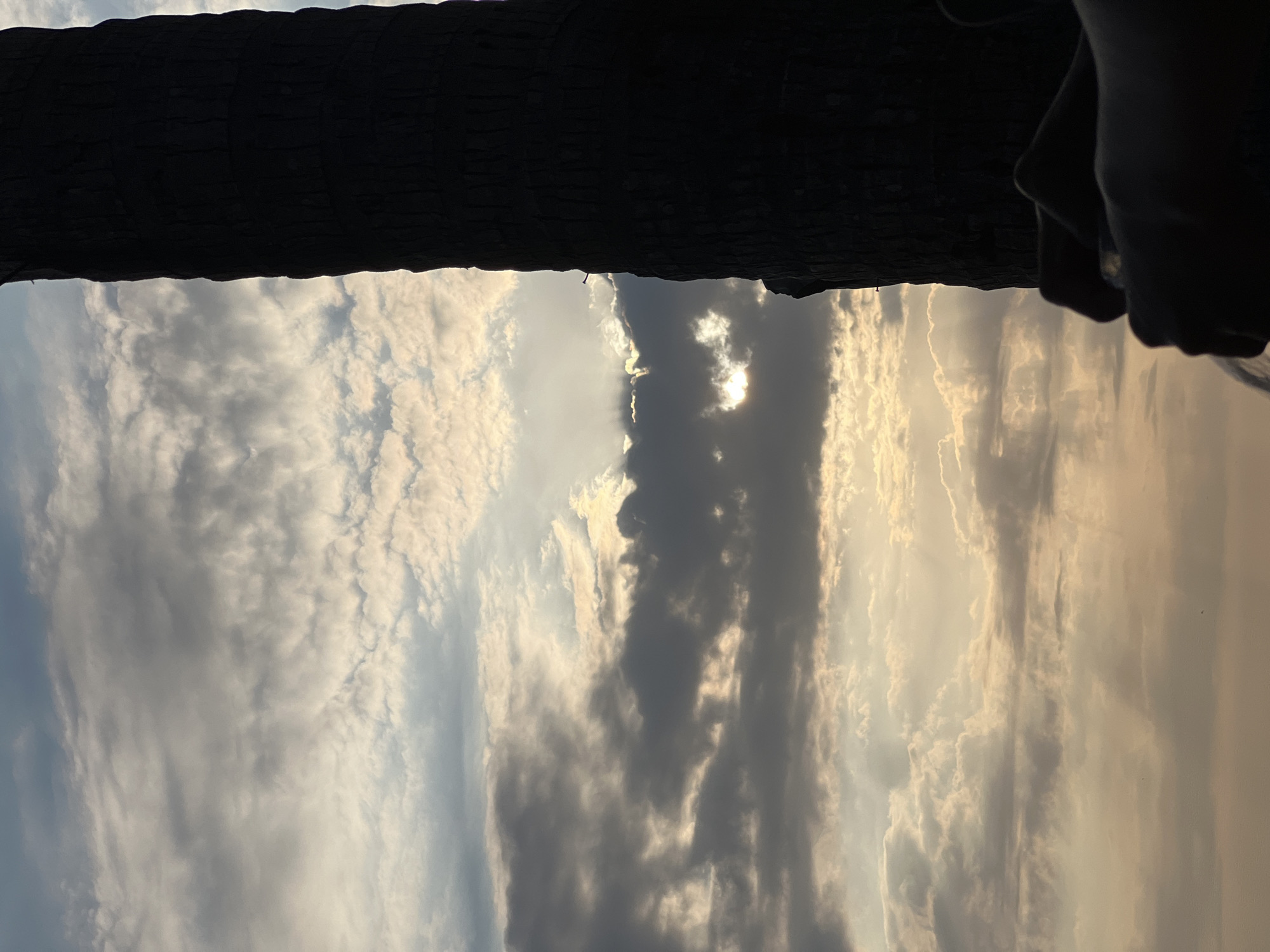 The image captures a close-up view of a person's hands holding a glass in silhouette against a dramatic sky. The sky is filled with a variety of clouds, with patches of sunlight breaking through, creating a dynamic and atmospheric effect. A dark, textured tree trunk dominates the right side of the frame, obscuring a portion of the sky. The setting appears to be outdoors, possibly a beachfront location in Kuta, Indonesia, given the tropical feel suggested by the palm tree and the overall lighting. The hands are holding a clear glass, which is slightly tilted, hinting at liquid being poured or consumed. The scene is lit by the warm, soft glow of what appears to be either sunset or sunrise.