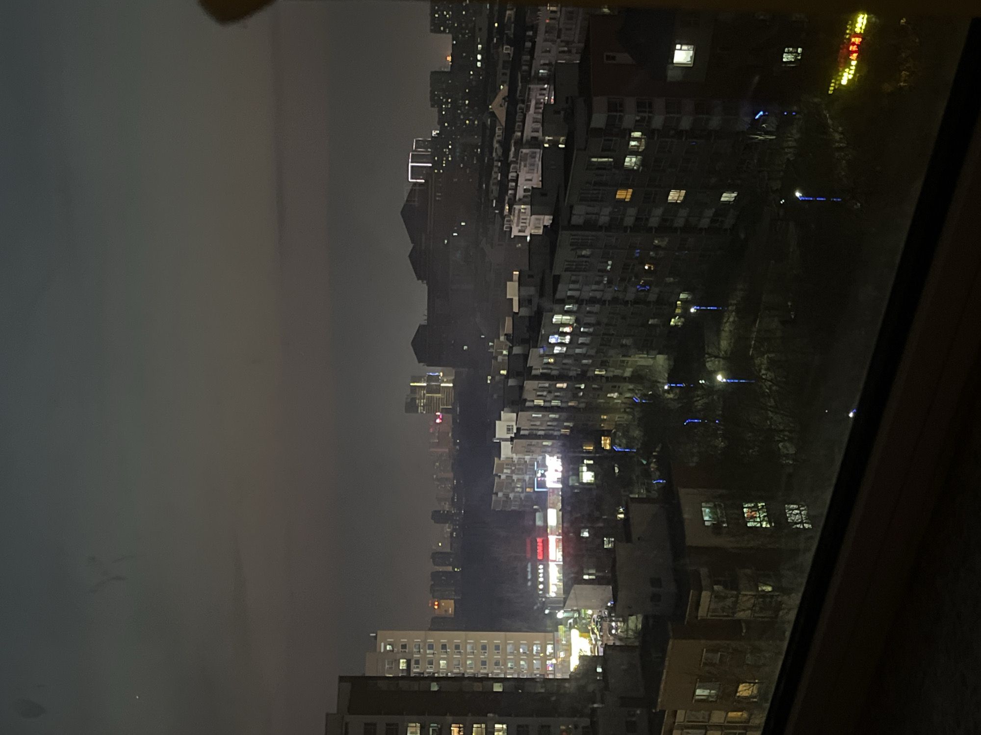 The image shows an elevated view of Changchun City, China at night. It's taken through a window, as indicated by the window frame on the right. The sky is overcast. The city's illuminated buildings and streets are visible. The cityscape comprises a mix of high-rise apartment blocks, commercial buildings, and other structures. Some buildings have brightly lit windows while others are dark. Streetlights and vehicle lights add to the city's illumination. In the foreground, there is a mix of residential and commercial buildings, including an apartment building, while in the distance more residential or commercial structures can be identified.