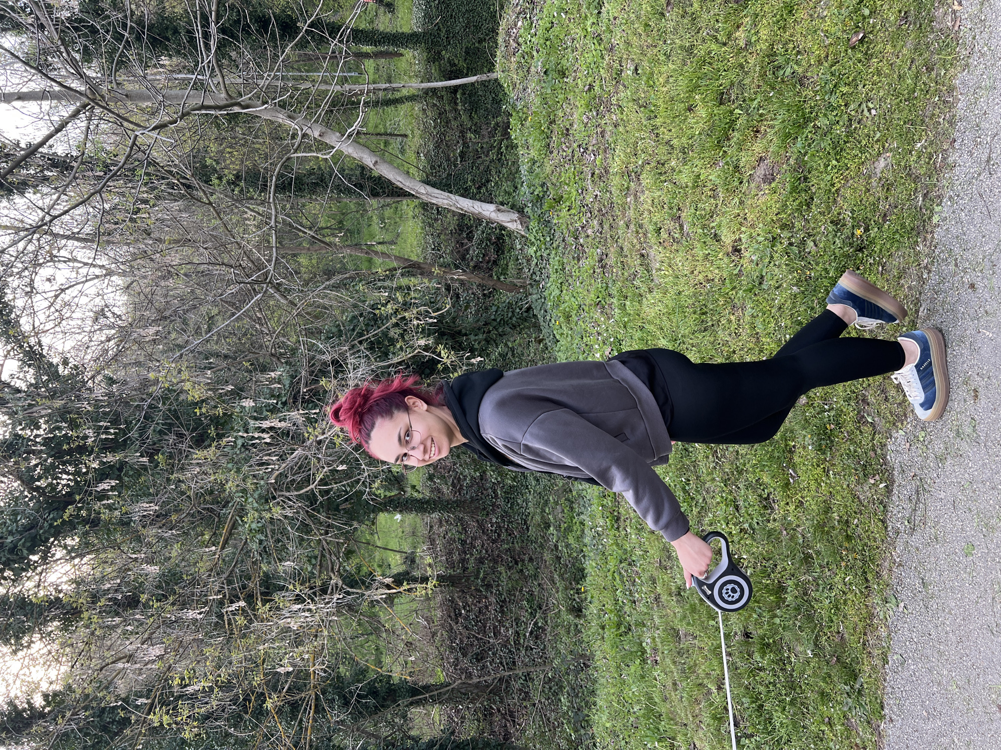 A young woman with bright red hair, tied back in a bun, stands smiling outdoors in what appears to be a park or green space. She is wearing glasses with thin frames, a two-tone grey and black hoodie, black leggings, and blue sneakers with thick, stacked light-colored soles (resembling Adidas Gazelle Bold). In her right hand, she holds a black and white retractable dog leash with a paw print design on its side, extending a white cord.
The setting features a paved path on the right side of the frame, bordering a grassy area to the left. The grass is vibrant green with some scattered weeds and small yellow flowers. In the midground and background, there are numerous deciduous trees, many with bare branches, indicating either late winter or early spring. Some trees show signs of budding with faint green leaves. Patches of dense green ivy cover parts of the ground and tree trunks further back. The lighting is soft and diffused, suggesting an overcast daytime sky. The woman's expression is pleasant and cheerful. The scene is located in Pleven, Bulgaria.