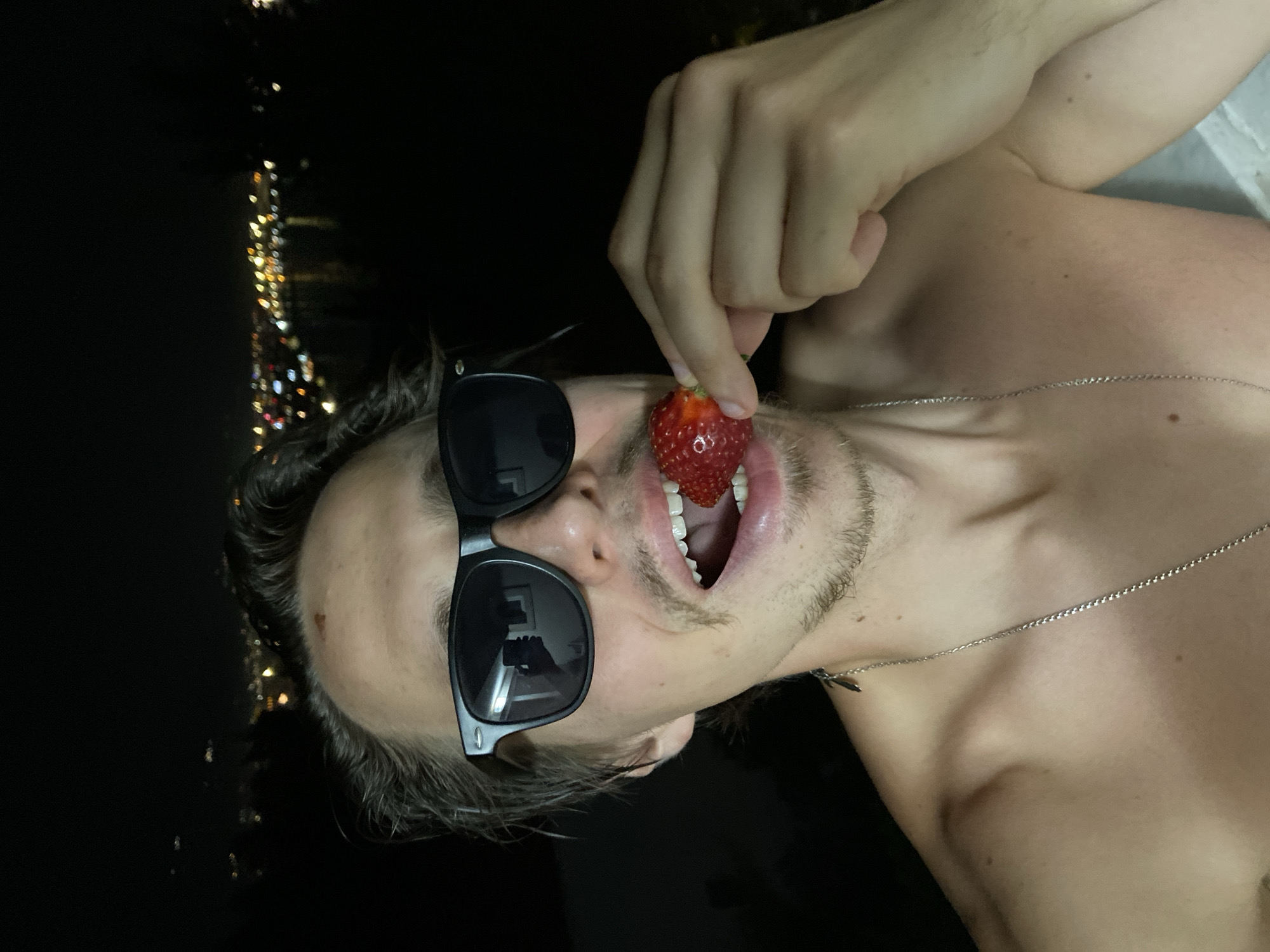 A shirtless man wearing sunglasses holds a strawberry in his mouth. He appears to be outdoors at night, with city lights visible in the background. The man is wearing a silver chain necklace. The city lights suggest a coastal location, potentially Šušanj, Montenegro.