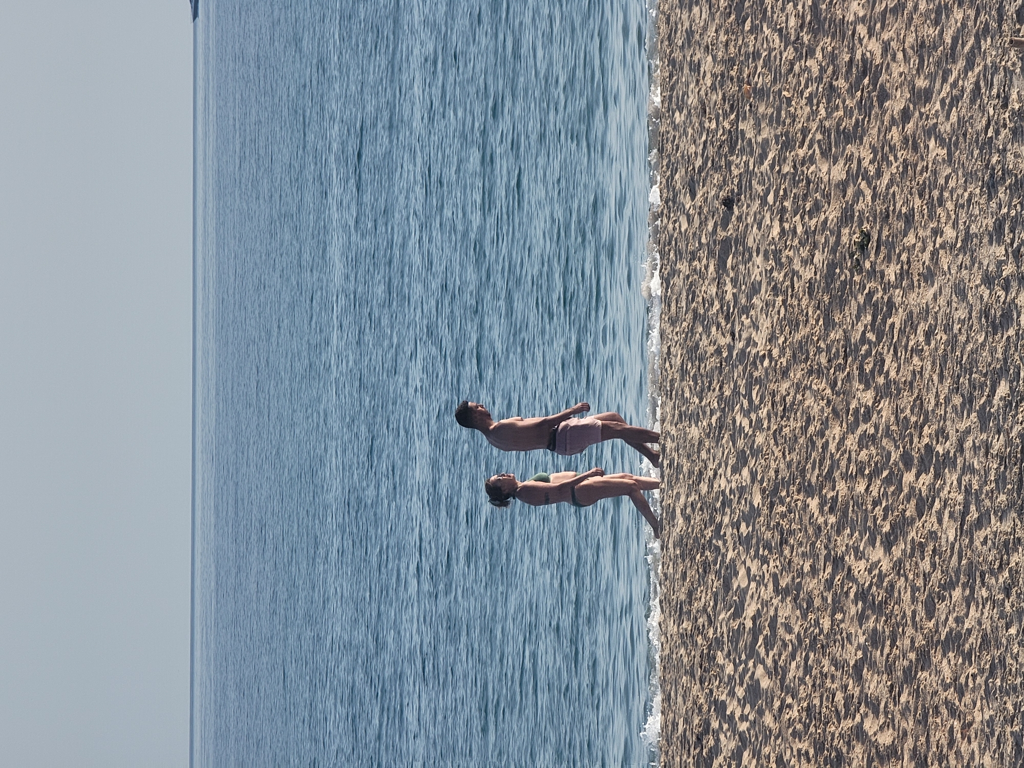 The image captures a serene scene on a beach in Quarteira, Portugal. The foreground is dominated by the textured sand of the beach, with gentle waves from the Atlantic Ocean lapping at the shore. In the mid-ground, a man and a woman, both in swimwear, walk along the water's edge, their figures silhouetted against the vast expanse of the sea. The ocean stretches out to the horizon, meeting a pale, cloudless sky. In the far distance, a solitary boat is visible, a small speck on the water. The overall impression is one of peacefulness and summer tranquility.