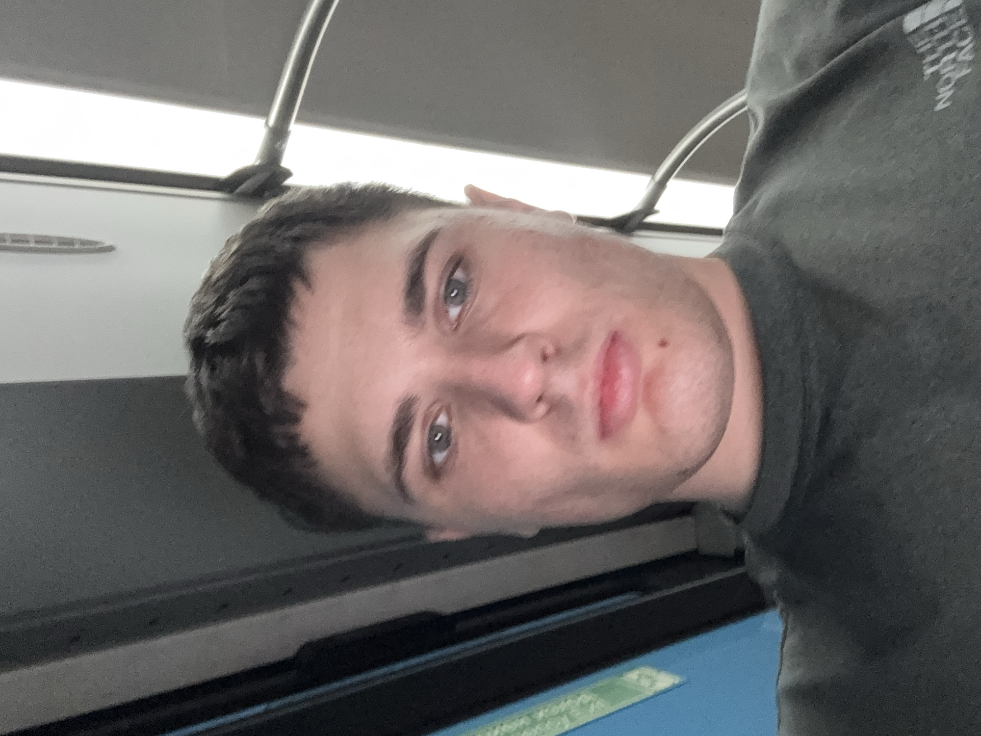A medium shot photograph, taken indoors with overhead lighting, shows a young Caucasian male with short brown hair and blue eyes. He is wearing a dark grey crewneck sweatshirt with a visible logo on the lower right side of the garment. The background consists of a gray wall, a metallic pole, and a bright light fixture. The lower left portion of the image reveals a blurred blue and green rectangular object. There is no indication of the location being in Stara Zagora, Bulgaria.