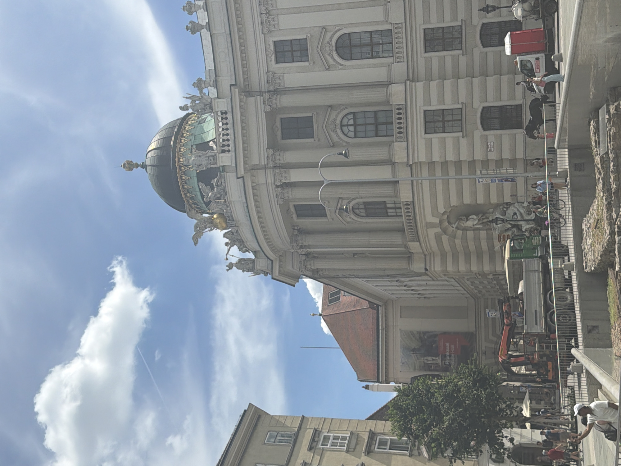 This media file captures an exterior view of a grand, ornate building with a dome, likely a historical or cultural landmark in Vienna, Austria. The building features classical architectural elements such as columns, arched windows, and intricate sculptures adorning its facade and dome. The sky above is a vibrant blue, dotted with fluffy white clouds, suggesting a bright and pleasant day. A contrail from an airplane is visible in the distance, hinting at the time of day being daytime.
In the foreground and mid-ground, several objects and people are present. A street lamp stands tall, its design fitting the classical aesthetic. Construction or maintenance work appears to be underway, indicated by a large truck with a crane and other vehicles. Several people are scattered around, some walking along the sidewalk, others seemingly engaged in conversation or observing the surroundings. Their attire suggests casual wear, suitable for a warm day. One person in the foreground is wearing a white cap and t-shirt, and is reaching down towards something on the ground, possibly interacting with another individual or an object.
A prominent red sign with white text is visible on a billboard-like structure further down the street. Although partially obscured, some text is legible, including "Festsaal" and "Staat". This suggests a public event or exhibition is taking place within the building. The presence of a large tree adds a touch of nature to the urban landscape. The overall scene conveys a sense of a bustling European city, with a blend of historical architecture and modern activity.