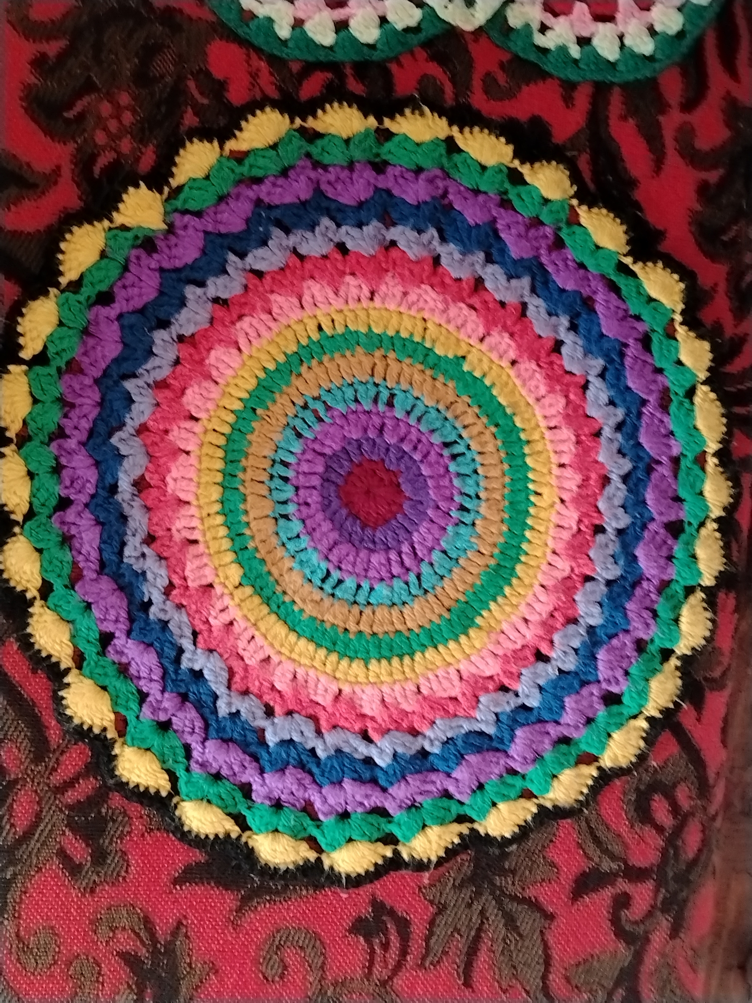 The image shows a colorful, crocheted circular mat or doily. The mat consists of concentric rings of various colors, including red, purple, light blue, tan, yellow, green, pink, blue, purple, and yellow. The background is a red and brown patterned fabric. There is no discernible information indicating the image location is in Яган, Russia.