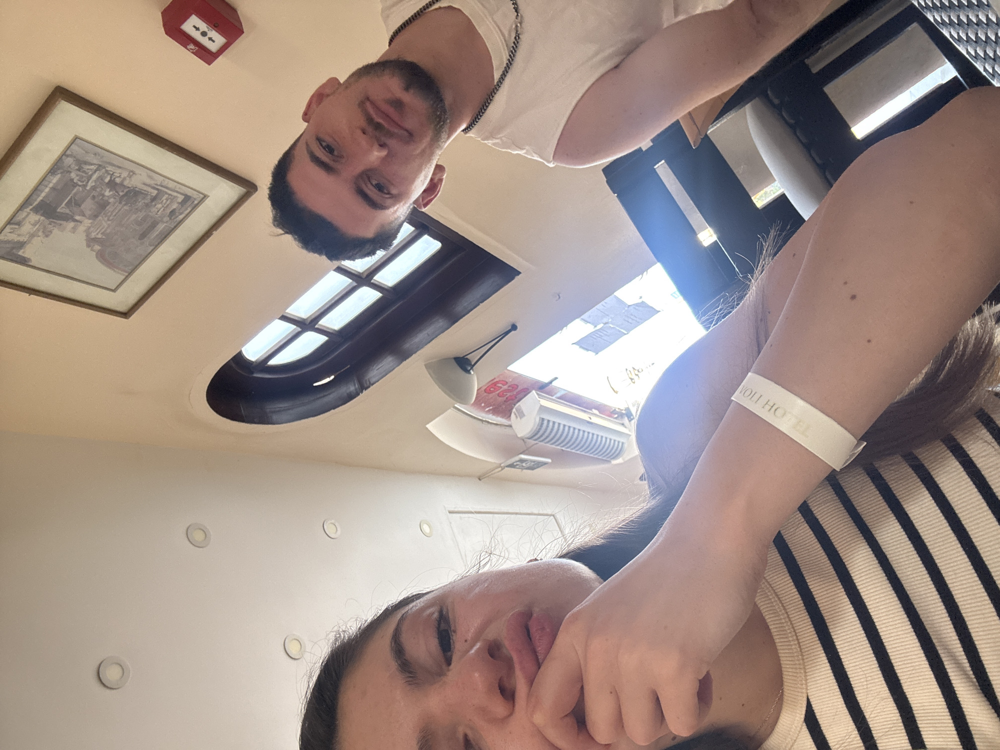 The image shows two individuals, a male and a female, in an interior setting. The male is positioned in the upper part of the frame, wearing a white T-shirt and a chain necklace. The female is in the lower part of the frame, wearing a striped shirt and a wristband that reads "Rivoli Hotel". An oval window is visible above them, with natural light streaming in. A picture frame and a red emergency device are mounted on the wall. The location is Unknown, Unknown.