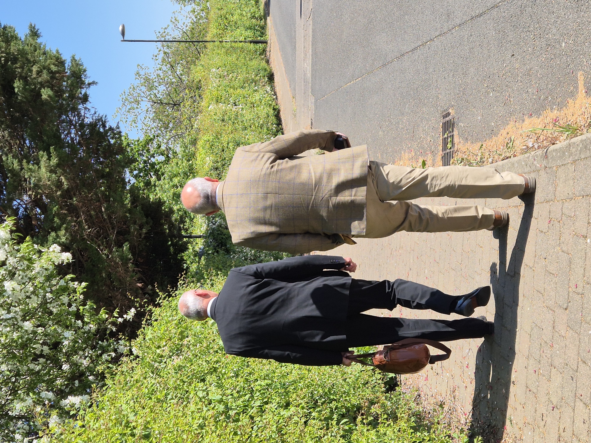 Two men are depicted walking away from the camera on a paved pathway under a clear blue sky in South Cambridgeshire, United Kingdom. The path runs alongside a strip of green foliage, including bushes with white blossoms and tall trees, and an asphalt road.
The man in the foreground, closer to the viewer, is dressed in a dark suit with a white shirt collar visible and is carrying a brown leather briefcase in his left hand. He has a bald head with grey hair on the sides. Slightly behind and to his right is another man, also with a bald head and grey hair, wearing a light brown plaid jacket and light-colored trousers.
The bright sunlight casts distinct shadows of the men on the path. A street light pole with a light fixture is visible against the clear sky in the upper background. Further down the asphalt road, a drain grate is noticeable. The abundant green vegetation and sunny conditions suggest a pleasant outdoor setting. No text is visible in the image.