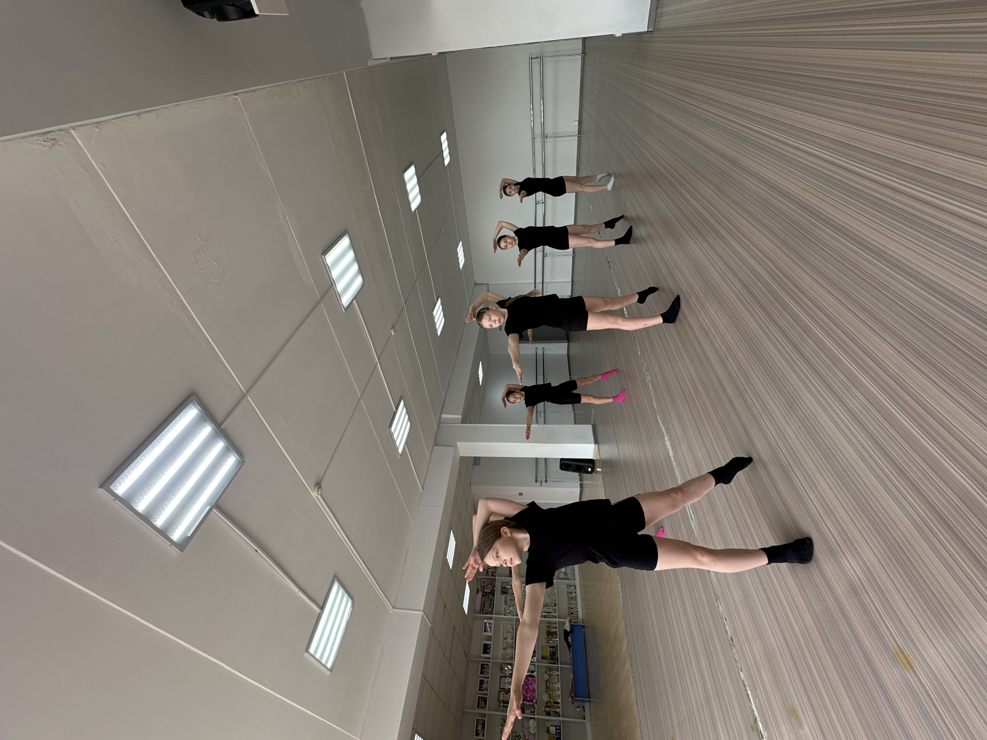 The scene is a dance studio in Votkinsk, Russia. Five young girls are performing a dance exercise. They are lying on their backs on a wood-look floor, with arms raised and one leg extended to the side. All are wearing black athletic attire. The room has a high ceiling with fluorescent lights. A mirrored wall is partially visible.