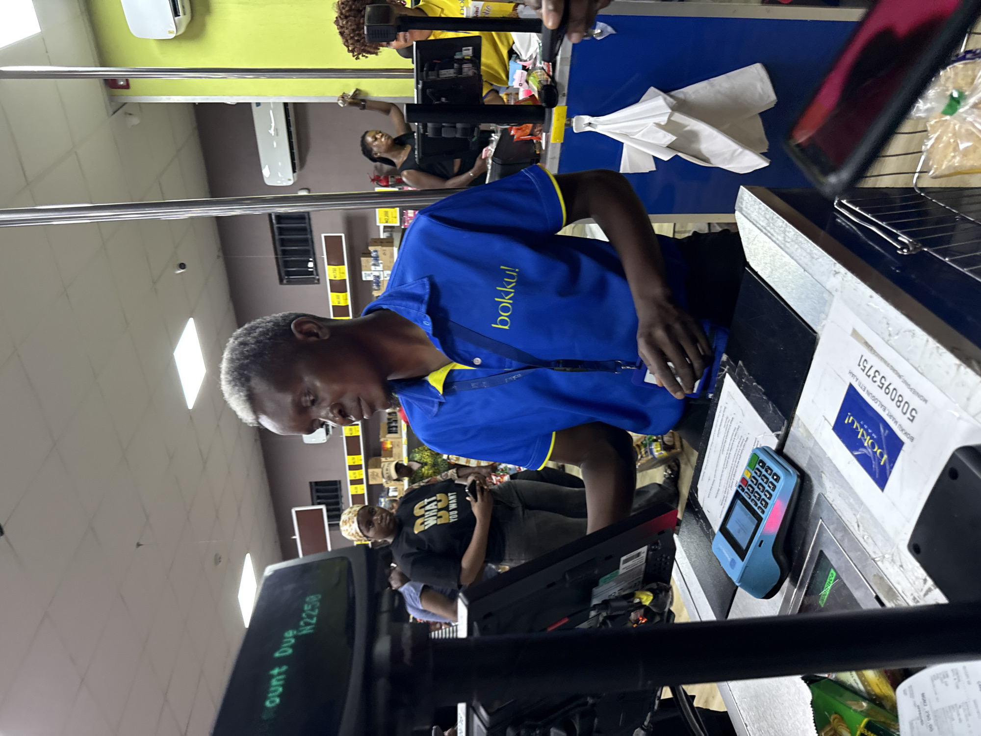 The image depicts a supermarket scene in Iranla, Nigeria, featuring a cashier wearing a blue "bokku!" uniform standing behind a checkout counter. The amount due is displayed as N2250. A customer is visible in front of the counter. Another customer, wearing a black t-shirt with the text "WHAT YOU WANT", is also present in the scene. On the counter, there is a receipt from "BOKKU MART, BALOGUN ETTI AJAH" with the number "5080953751", along with a point-of-sale (POS) device. Other objects include packaged food, a metal divider, and additional white receipts or paper. The supermarket's lighting is provided by overhead fluorescent lights and air conditioning units are mounted high on the walls. The environment appears to be busy, with other customers and supermarket shelving partially visible in the background.