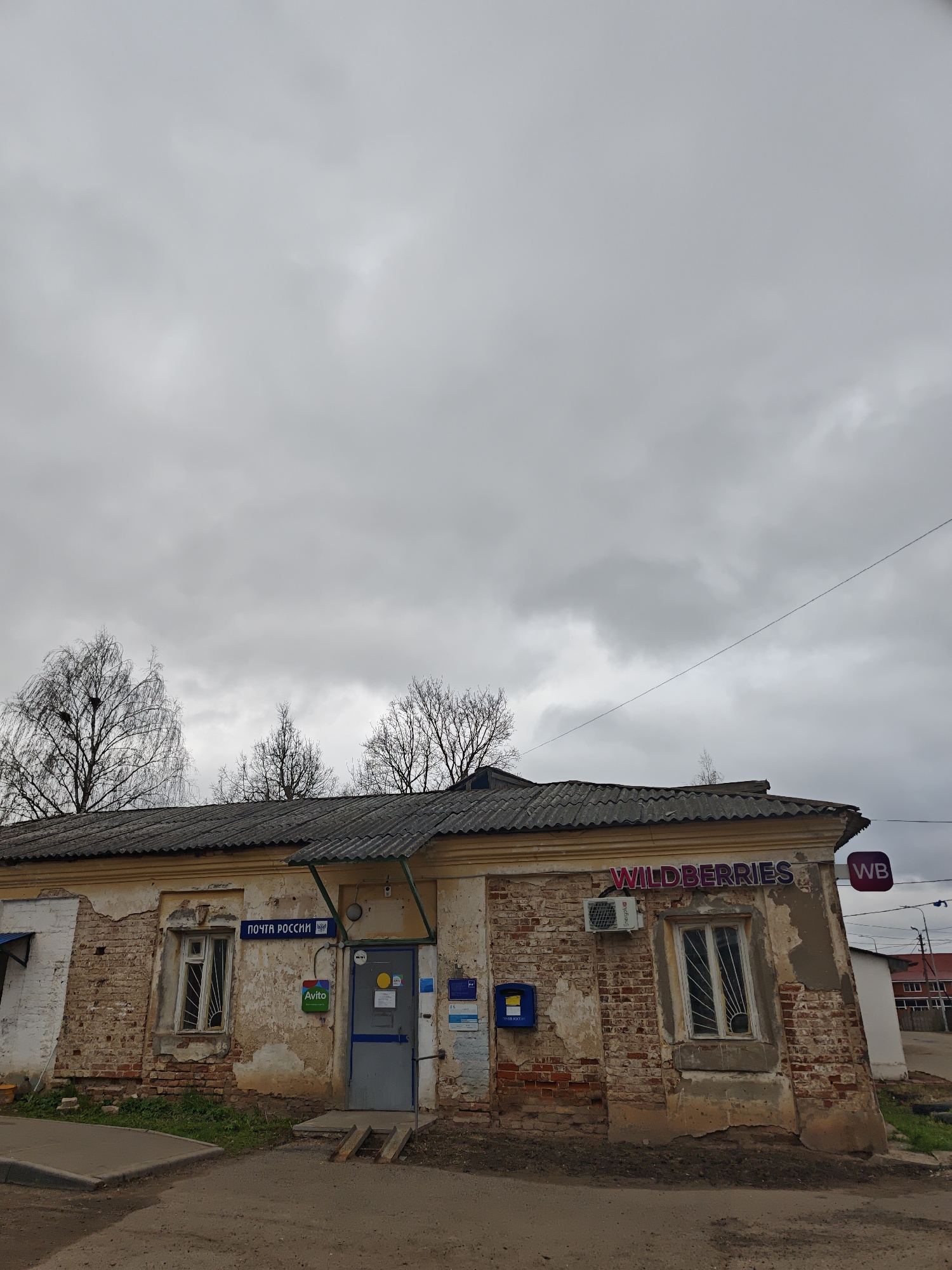 The image captures a dilapidated building in посёлок совхоза "Буденновец", Russia. The structure appears to house both a почта россии (Russian Post Office) and a Wildberries location, indicated by signage. The building is constructed primarily of brick, with extensive crumbling and deterioration of the facade, particularly the plaster. The roof is tiled and exhibits some sagging. Windows are present on both sides of the entrance, covered with bars. A blue mailbox is visible. An Avito sign is next to the entrance. The sky is overcast, and there are bare trees in the background. The area in front of the building appears to be a paved road or parking area. The overall impression is one of neglect and disrepair.