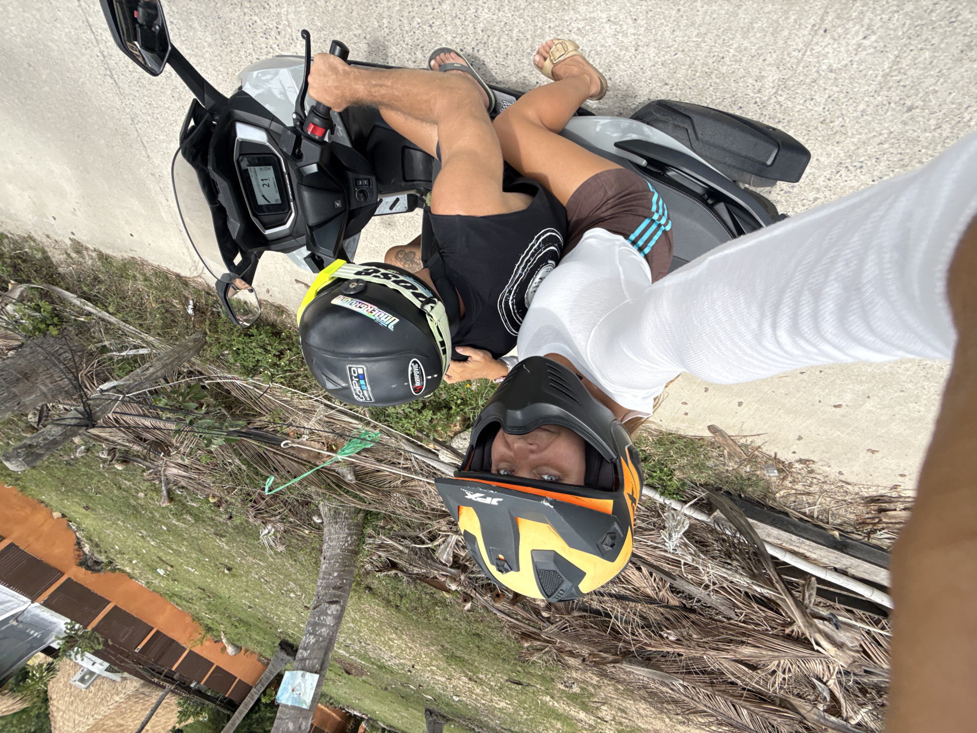 In Ko Samui District, Thailand, two individuals are riding a scooter on a paved road bordered by sparse vegetation and dried palm fronds. The person in the front, wearing a white shirt and an orange and black helmet, is looking towards the camera. The person in the back, wearing a black t-shirt and a black and yellow helmet, is operating the scooter. The scooter's digital display shows "21". In the background, there is a building with an orange-colored wall and a thatched roof structure. A portion of a palm tree trunk is also visible.