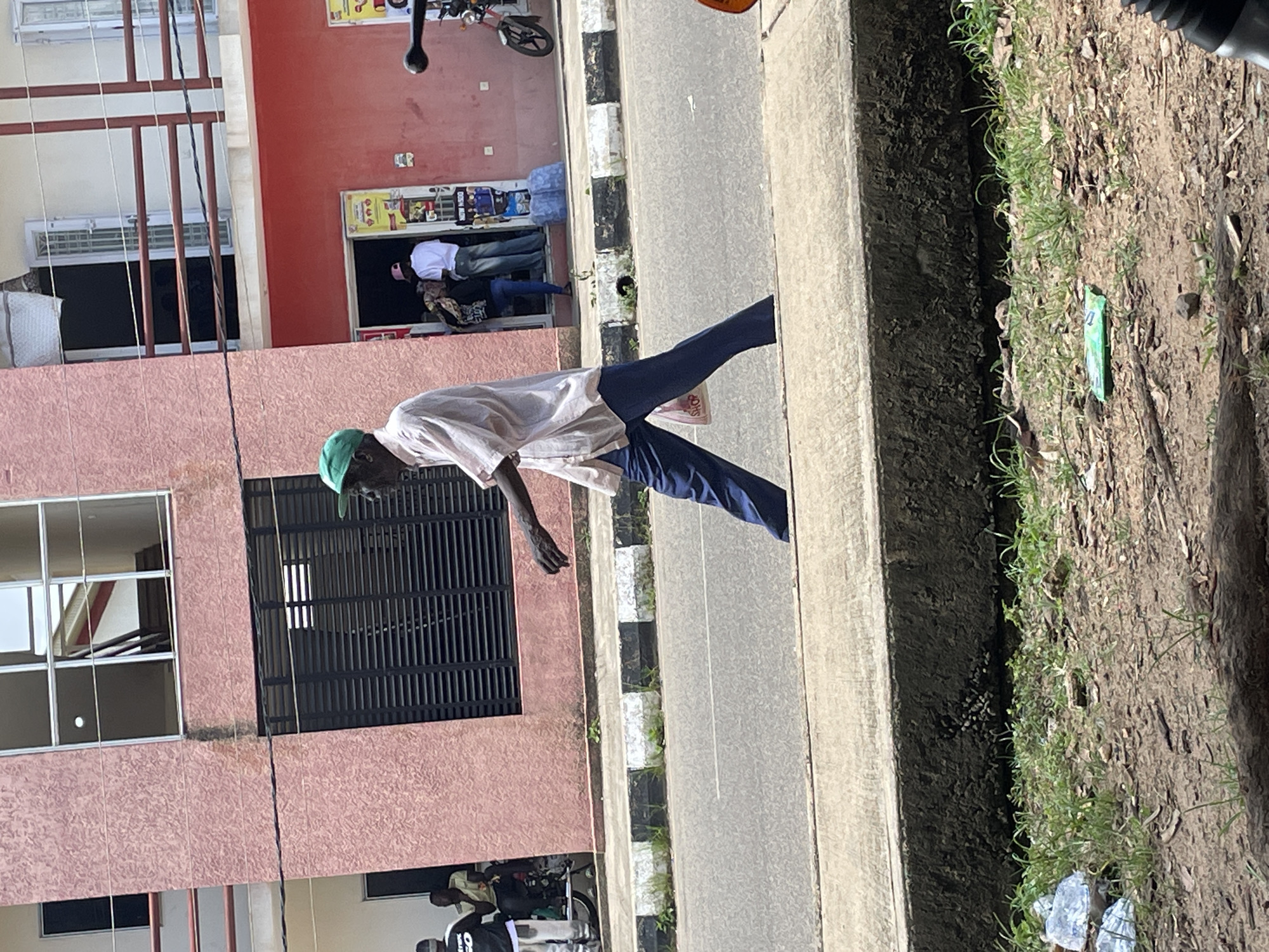 A man wearing a light-colored long-sleeved shirt, dark blue pants, and a green cap walks across a paved street in front of a pink building. He is holding a white plastic bag in his left hand. In the background, the pink building has multiple windows and balconies. A red door on the building leads to a shop. To the right of the shop, a red motorcycle is parked. In the far background, there are other buildings. The street has a raised concrete sidewalk. The ground in the foreground is covered with dirt, grass, and debris. The scene appears to be an urban environment in Jos, Nigeria.