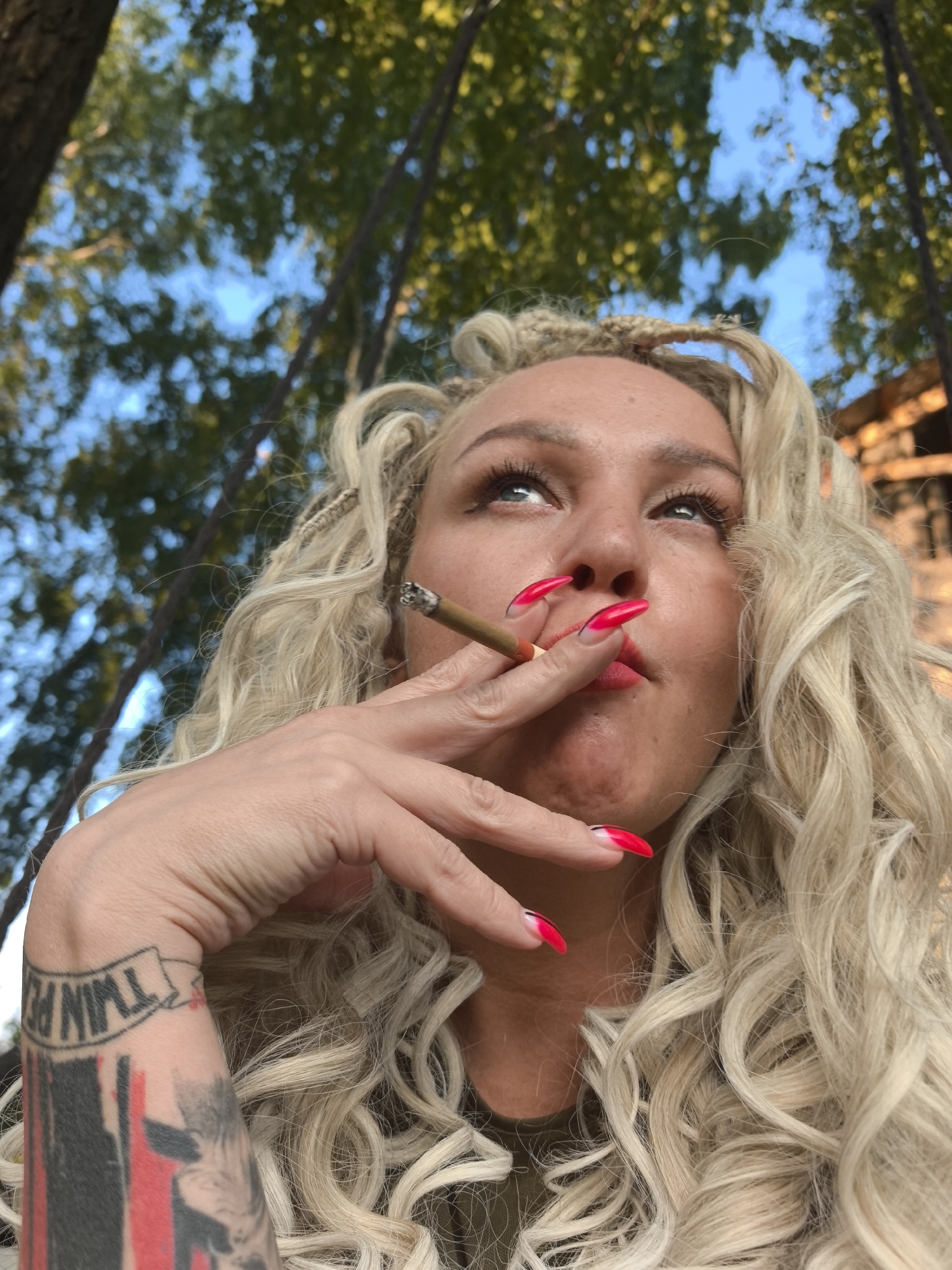 A medium shot from a low angle shows a woman with blonde curly hair and braided sections smoking a cigarette outdoors, with trees and the sky in the background. She is looking up and to her right. The woman has bright red nails with white tips and a tattoo on her left forearm. The overall lighting suggests it is daytime. The image does not contain information to determine the location in the city of Noviy, Russia.