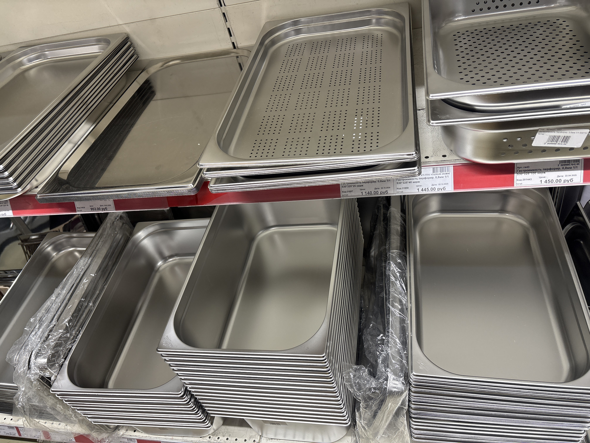 The image shows a retail display of stainless steel gastro-norm trays, likely in a restaurant supply store. The trays are stacked on shelves, organized by size and type, some with perforations. Price tags with Russian currency (rubles) are visible. The tags suggest that the items were photographed in Krasnoyarsk, Russia as of at least October 2025.