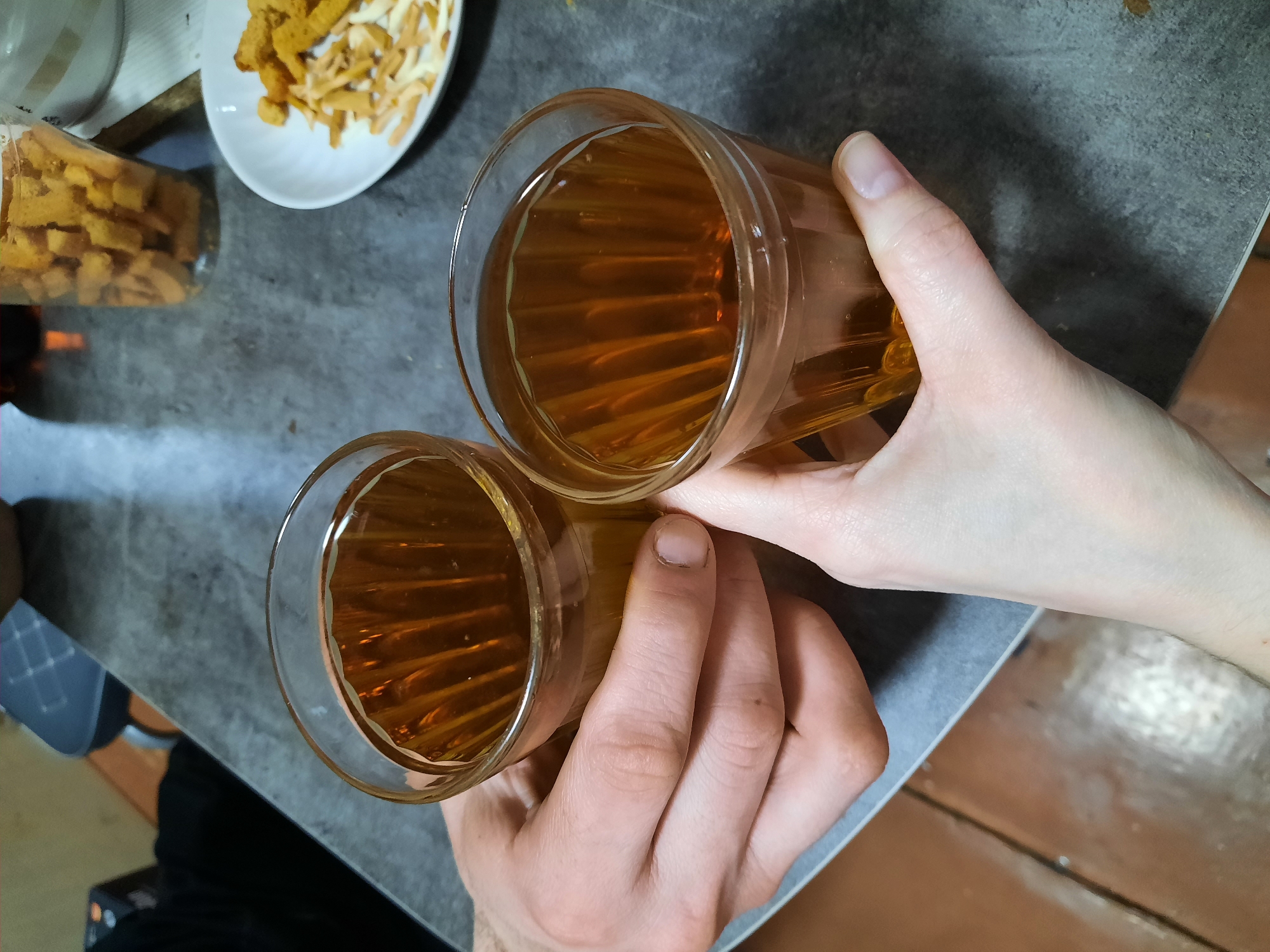 Two individuals are holding clear, faceted glass tumblers containing an amber-colored liquid, positioned above a gray, speckled tabletop in the city of Krasnogorskoye, Russia. A male hand, distinguished by its larger size and slightly darker skin tone, grips the left glass. A female hand, characterized by its lighter skin tone and manicured nails, holds the right glass. Both hands are in close proximity, suggesting an interaction such as a toast or shared drink. In the upper right background, a white plate holds an assortment of dry snacks, including small rectangular croutons and thin, lighter-colored strips. Behind the plate, a clear jar filled with similar rectangular croutons is visible. The immediate background shows an out-of-focus dark object and a reddish-brown tiled floor.