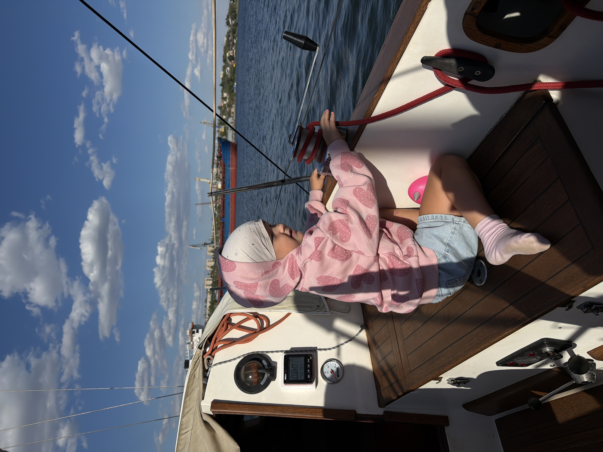 A young girl wearing a pink hooded sweatshirt with a strawberry pattern and light blue denim shorts is seated on the deck of a sailboat. She has a light-colored headscarf on. She is holding a red rope which is wrapped around a winch. The boat has wooden decks and white siding. There are several gauges and instruments mounted on the interior wall of the boat. The sailboat is on a body of water, likely a river or bay, with a large cargo ship and industrial cranes visible in the background on the shore. The sky is blue with scattered white clouds, indicating a sunny day. The scene appears to be taking place in the city of Kamyshin, Russia, given the presence of industrial port infrastructure that is consistent with the appearance of the Kamyshin port on the Volga River.