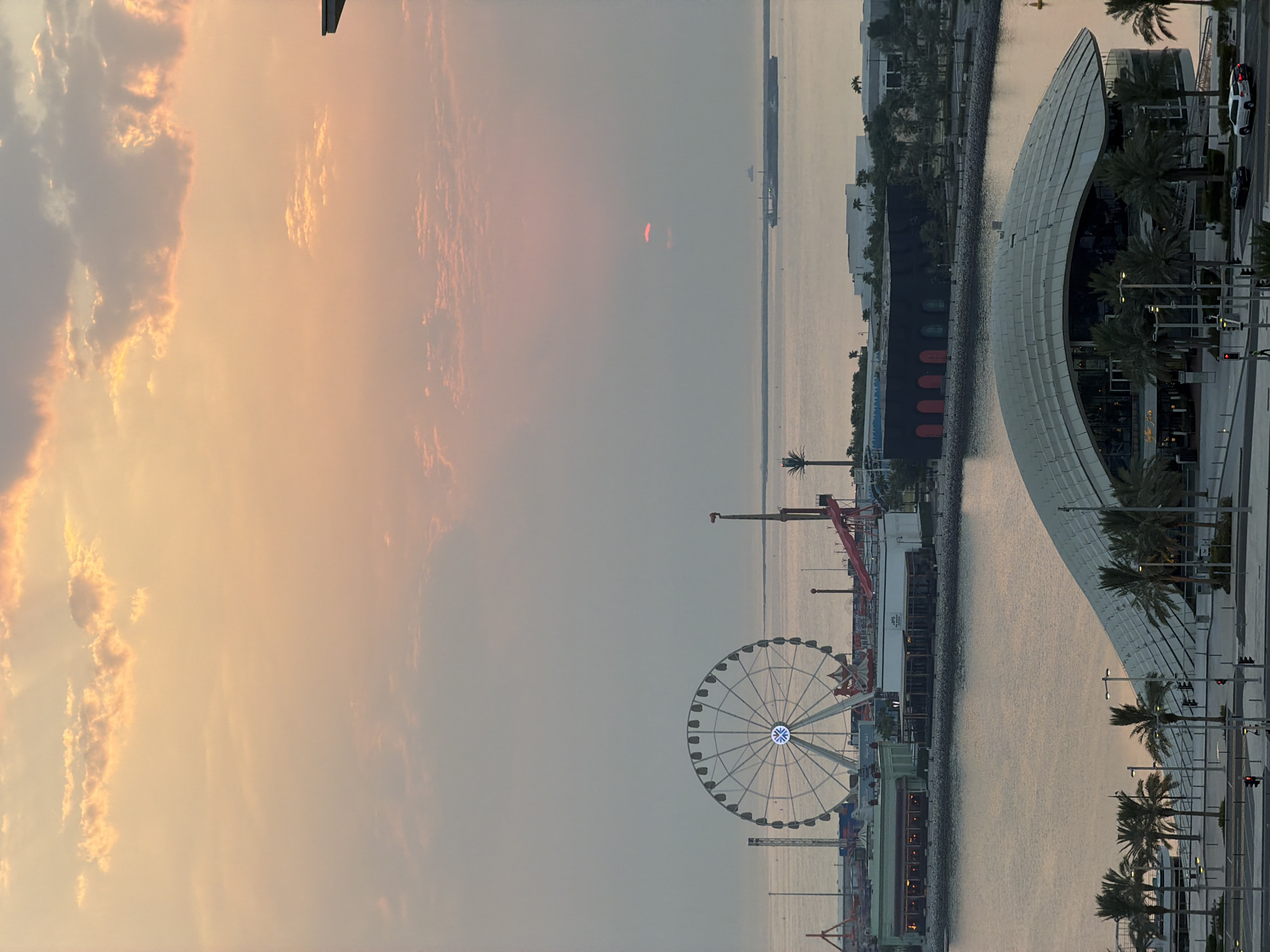 The image captures a panoramic view of the Lusail City waterfront at dusk. The sky is a soft gradient of orange and pink, with scattered clouds catching the fading sunlight. A large Ferris wheel stands prominently on the left side of the frame, illuminated with white lights, suggesting an amusement park or entertainment district. Several other amusement park rides and structures are visible near the Ferris wheel.
In the middle ground, a body of water separates the amusement area from a strip of buildings lining the shore. These buildings appear to be restaurants or commercial establishments, with some showing interior lights. Further in the distance, a large ship is visible on the horizon.
In the foreground, a modern, curved-roof building with a glass facade dominates the scene. Palm trees line the walkway in front of this building, indicating a tropical or subtropical climate. Streets with traffic lights and a few cars are visible, suggesting active urban infrastructure. The overall impression is one of a vibrant, modern city by the sea, bathed in the warm hues of sunset.