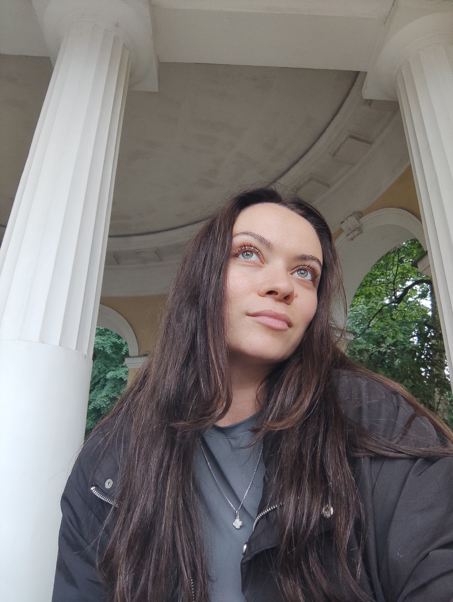 A woman with long, dark brown hair is captured in a close-up, low-angle shot from the chest up, looking upwards and to the right. She is wearing a dark jacket over a gray t-shirt with a delicate silver pendant necklace. Her facial features are detailed, with striking blue eyes, light eyebrows, and fair skin. The background reveals architectural elements, specifically the fluted white columns and arched openings of a building that appears to be in a park setting with lush green trees visible through the arches. This setting is characteristic of a neoclassical or baroque structure, often found in St. Petersburg, Russia's imperial palaces and gardens, such as those in Tsarskoye Selo or Peterhof. The overall lighting is soft and diffused, suggesting an overcast day or shaded area.