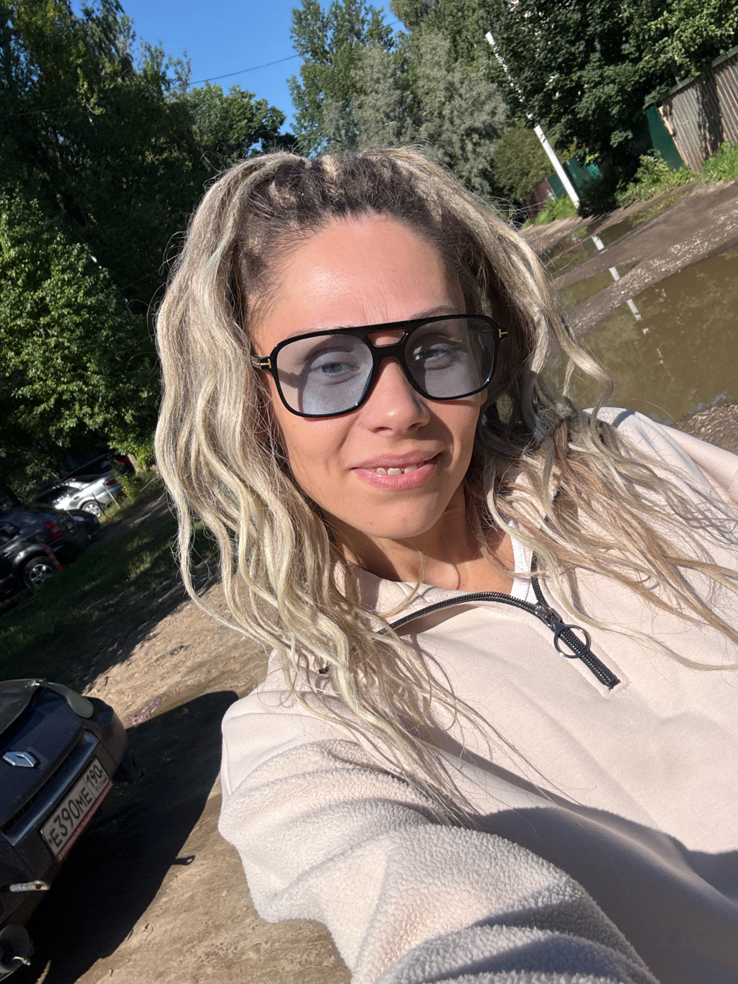 In Rusanovo, Russia, a blonde woman with braided hair and sunglasses is taking a selfie outdoors. She is wearing a light-colored hooded sweatshirt with a zipper. The background shows trees, a dirt road, and a puddle of water. A dark-colored car is parked on the left side of the image. The overall lighting suggests a sunny day.