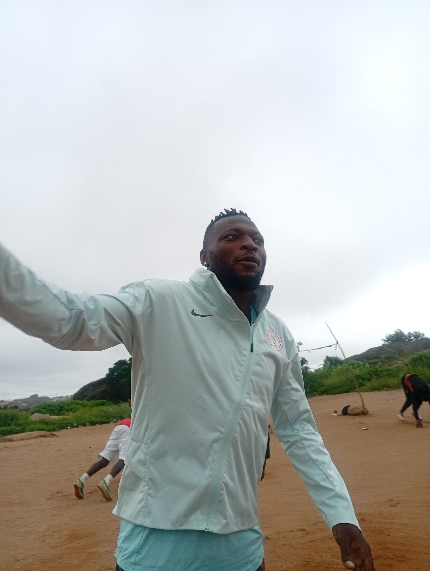 A man, wearing a light blue Nike jacket over a teal shirt and blue shorts, stands in the foreground, his left arm outstretched as if addressing someone. He has short, dark, braided hair, a beard, and is wearing earrings. Behind him, a person in a red shirt and white shorts is doing push-ups on the sandy ground. Further in the background, another individual in black pants and a red shirt with black accents is walking. The environment is an outdoor, sandy area with sparse vegetation and rocky outcrops in the distance under an overcast sky. This scene is likely located in Jos, Nigeria, a city known for its elevated terrain and rocky landscapes.