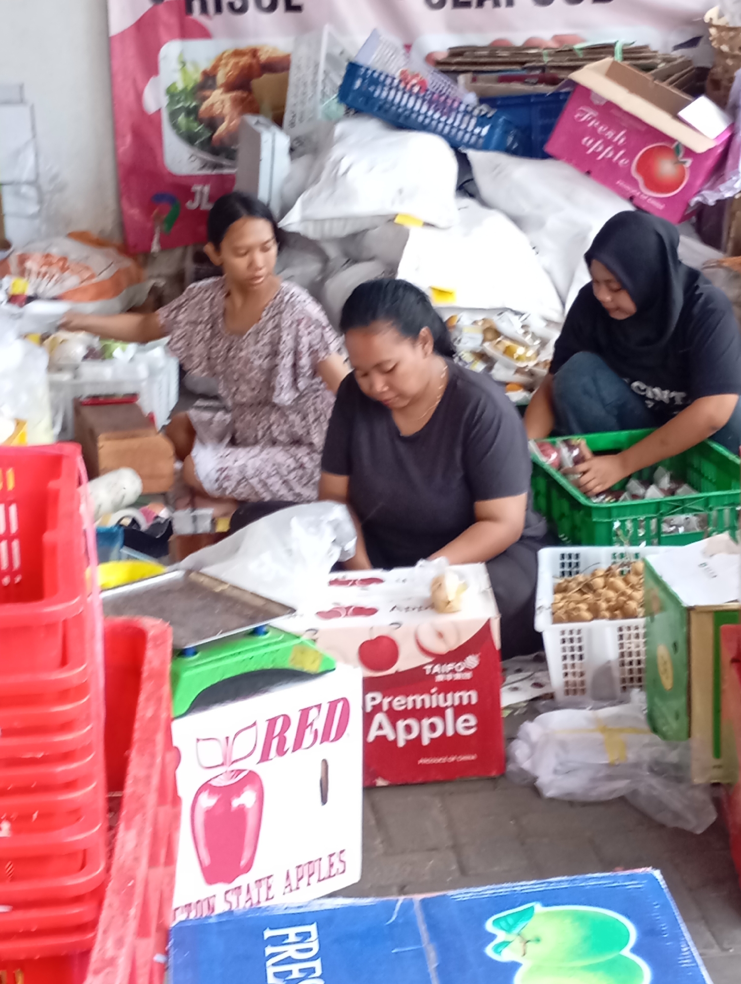 In Plintahan, Indonesia, three women are engaged in sorting and packaging produce, likely at a market or distribution center. The scene is cluttered with various containers and goods. In the foreground, to the left, are stacked red plastic crates. In the center foreground, a white box with an image of red apples and the text "RED" and "STATE APPLES" is visible. Another box to its right also features an apple graphic and "Premium Apple".
Two women are seated on the ground. The woman on the left, wearing a patterned dress, appears to be working with smaller items on a surface in front of her. The woman in the center, in a dark shirt, is looking down at the box with "Premium Apple". A third woman, to the right, wearing a hijab and dark shirt, is crouched down and sorting items within a green plastic crate. In the background, there are large white sacks, cardboard boxes, and other crates, suggesting an active commercial environment. The ground appears to be paved with cobblestones.