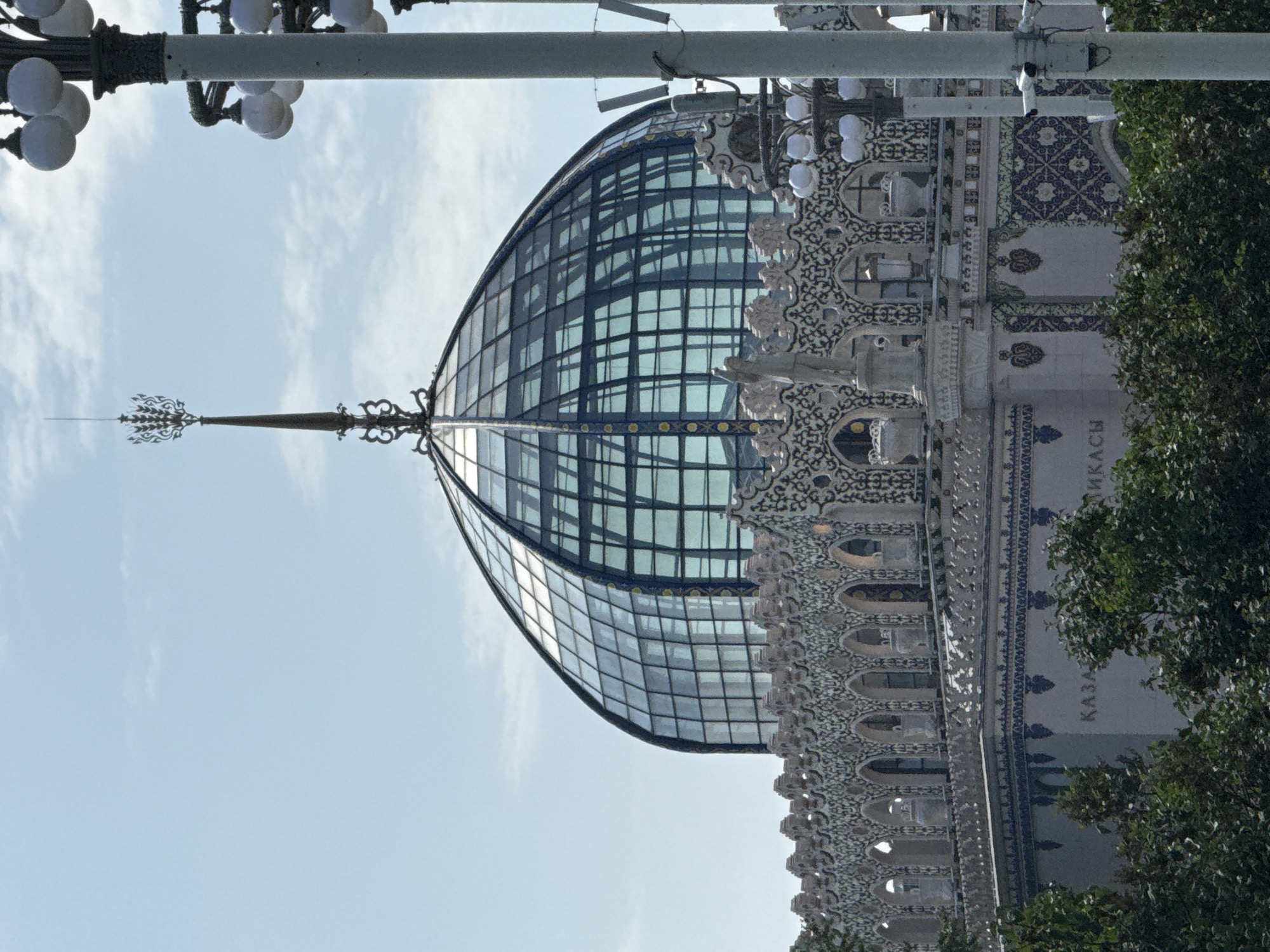 The image shows the pavilion of the Tatar ASSR (now the Republic of Tatarstan) at VDNKh (Exhibition of Achievements of National Economy) in Moscow, Russia. The pavilion features a prominent glass dome with a golden spire. The facade is decorated with intricate mosaic patterns and architectural details, including arched windows and sculptures. Several streetlights with multiple globe-shaped lamps are visible to the right of the pavilion, and lush green trees partially obscure the lower portion of the building. The sky above is clear with scattered clouds. The word "KAZAN" (referring to the capital of Tatarstan) is visible in raised letters on the building.