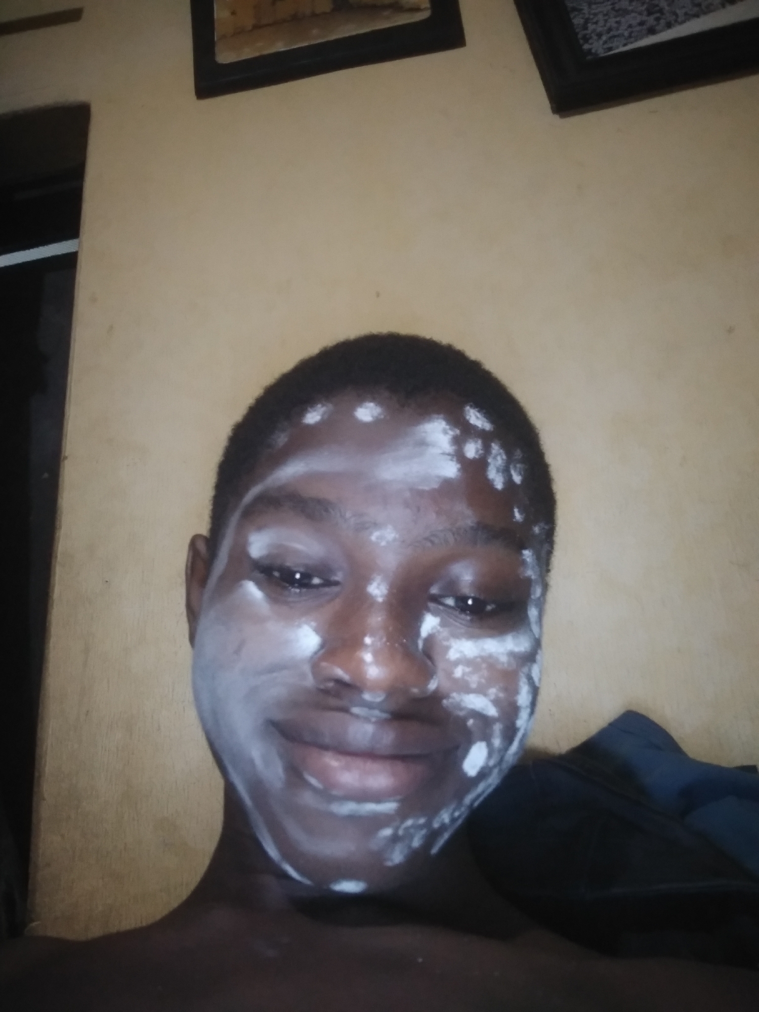 A close-up, indoor photograph captures a dark-skinned person with white substance applied to their face. The substance appears to be applied unevenly. Two framed images hang on the wall behind the person. A dark, partially visible object is present to the person's right. The location is inferred to be Shangisha, Nigeria, based on the prompt's mention of the location. No specific actions, interactions or objects related to that location are observable.