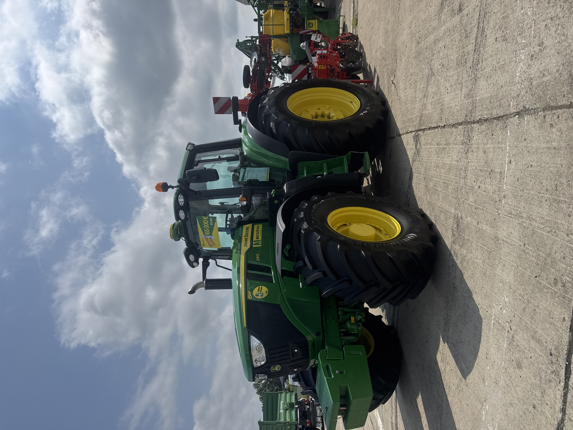 The image, originally rotated, captures a green and yellow John Deere tractor prominently displayed on a concrete surface under a partly cloudy sky. The scene is likely an outdoor dealership lot or agricultural exhibition in Stara Zagora, Bulgaria.
The tractor features the "JOHN DEERE" logo on its hood and "МЕГАТРОН" (Megatron), a common agricultural machinery dealer in Bulgaria, on its side. Below the "6M" model designation, a "240" is visible, along with a circular decal featuring a large "5", possibly indicating a warranty or special offer. A yellow sign affixed to the front windshield displays "ИПОЛО ПРЕДЛАГА" (Hypolo Offers) and a price of "165 000€ БЕЗ ДДС" (165,000 Euros without VAT), suggesting the tractor is for sale.
In the background, other agricultural equipment is partially visible, including what appears to be a planter or sprayer with yellow tanks, and a green trailer that shows "BRANTH" text on its side. The concrete ground extends across the foreground and mid-ground. The bright, diffuse lighting and subtle shadows indicate it is daytime. No people are present in the image; the focus is solely on the agricultural machinery.