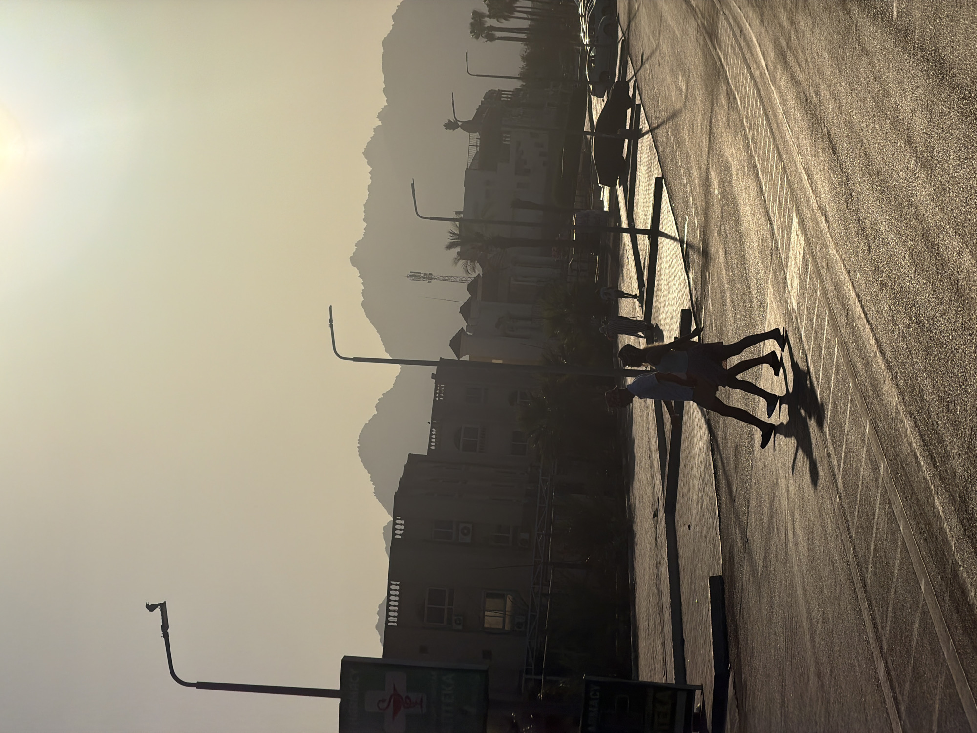 In Sharm El Sheikh, Egypt, a pedestrian crossing is depicted during a sunset with the sun low in the sky, casting long shadows. Two individuals, silhouetted against the light, are walking across the street. A streetlamp stands to the left, and further in the background, a line of buildings and palm trees are visible, with mountains forming a hazy silhouette against the bright sky. The street is wet, reflecting the sunlight. A sign for a pharmacy is partially visible on the left. Cars are parked in a lot in the mid-ground.