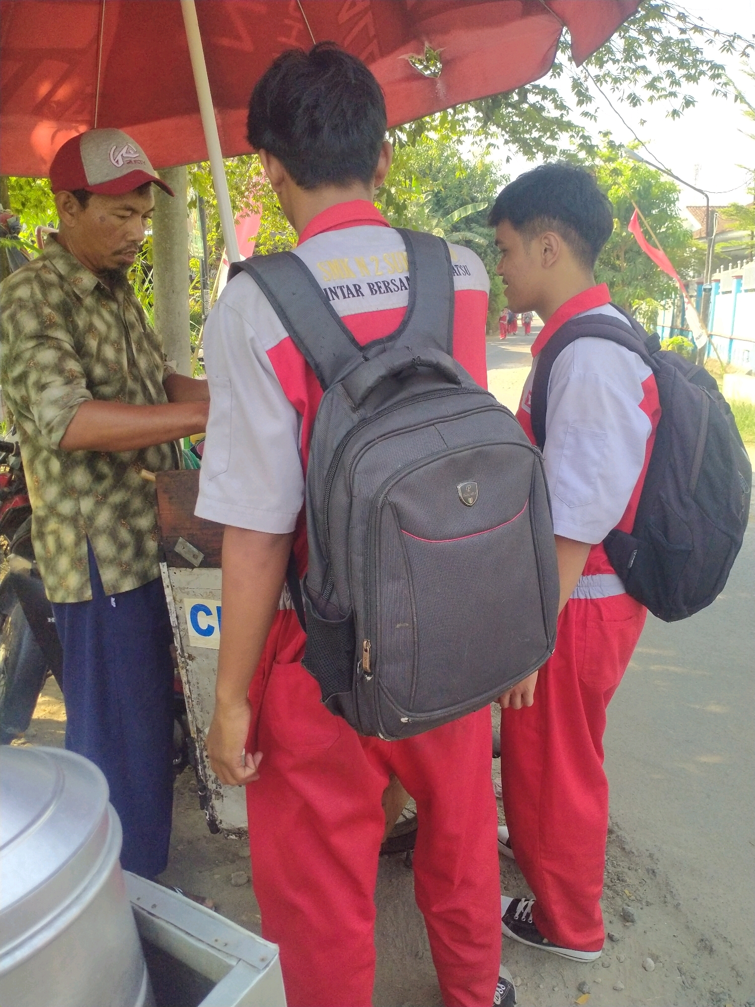 In Begajah, Indonesia, a man wearing a patterned shirt and a baseball cap stands under a red umbrella. He appears to be interacting with two young individuals, both wearing red and white school uniforms and backpacks. One of the students is facing away from the camera, while the other is partially visible from the side. A motorcycle is parked behind the man. In the background, there are trees, a white fence, and a building. The ground is a dirt and asphalt road.