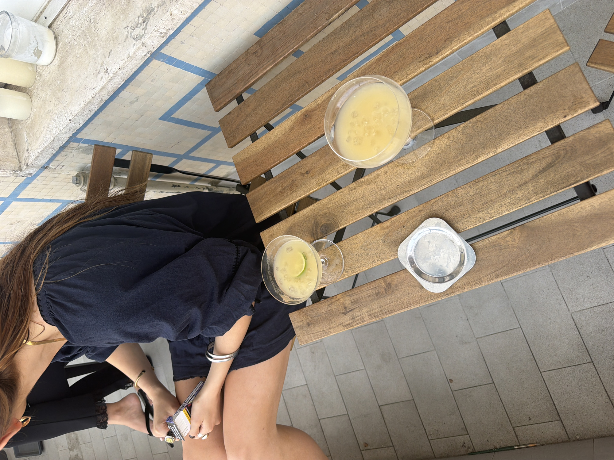 The image captures an overhead view of a person seated at a wooden table on a terrace. The person is wearing a dark blue, loose-fitting top and dark pants. They are holding a small object, possibly a lighter or a pack of cigarettes, in their hands.
On the table, there are two martini glasses filled with a yellow liquid, one with a lime slice. There is also a small, square metal coaster. The table is made of wooden slats with metal supports. The floor surrounding the table is paved with large, light grey tiles. In the top left corner, there are white cylindrical objects, possibly candles or decorative containers, positioned near a wall with patterned tiles.
Without additional context, determining the precise location in Milan, Italy is not possible.