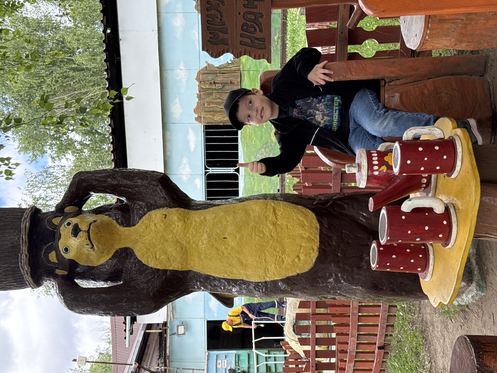 In the city of Chaikovsky, Russia, a young boy wearing a black cap and jacket over a t-shirt with a dinosaur graphic, and blue jeans, sits on a wooden chair in an outdoor setting. He is giving a thumbs-up gesture. In front of him, on a yellow table supported by a tree stump, are three red and white polka-dotted mugs and a red teapot. To the left of the boy, a large, brown, bear-shaped statue with a yellow body stands next to the table. Behind the statue and boy, a building with a light blue facade is visible, with a painted landscape of mountains and sky on its side. A red wooden fence surrounds the area. In the background, a worker in a yellow hat and blue overalls is seen near a doorway. The environment appears to be an amusement park or children's play area.