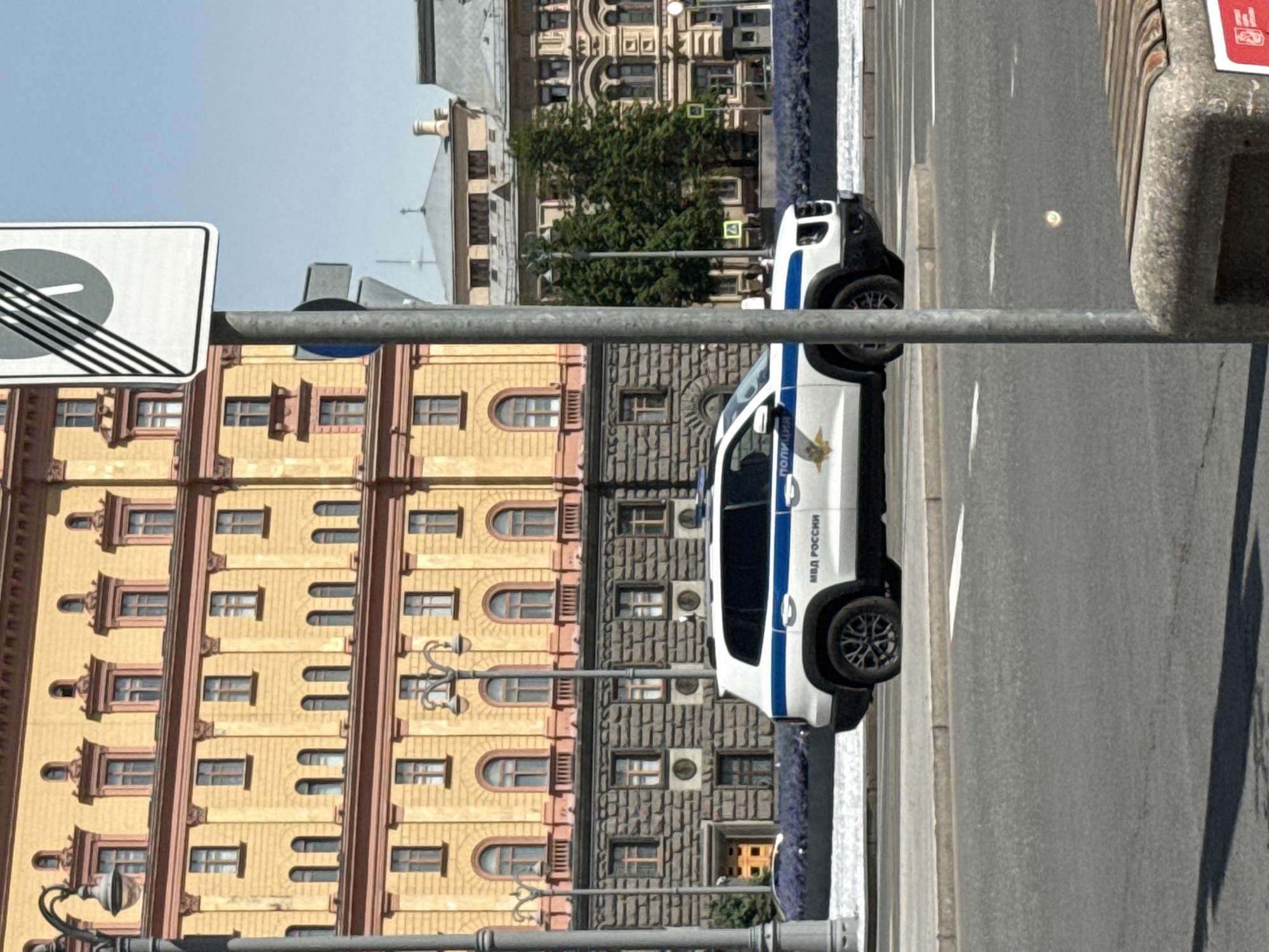 The image shows a street in Moscow, Russia. A white police car with blue stripes and the inscription "МВД РОССИИ" (MVD ROSSII) is parked on the side of the road. Behind the car, there are buildings with classical architecture. A street sign depicting a gray circle with black diagonal lines across it is on a pole in the foreground.