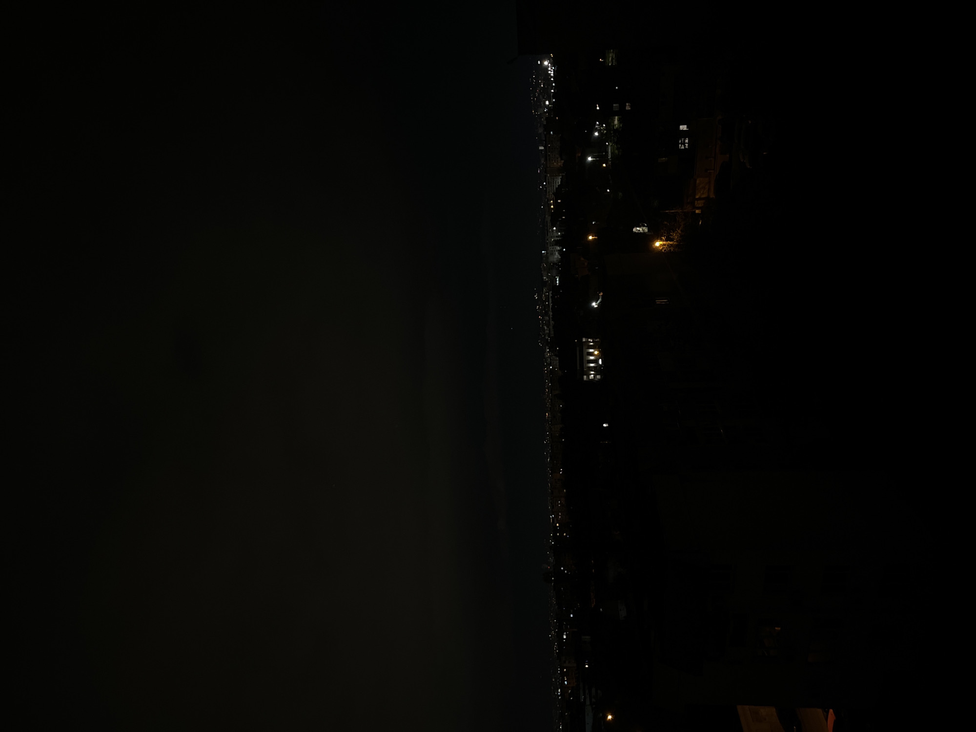 The image depicts a nighttime view of a coastline. On the right side, there is a dark expanse suggestive of land with scattered light sources that appear to be buildings and streetlights. The lights are arranged in lines, indicating roads or streets. The left side of the image is mostly dark and uniform, likely representing water. The specific location within Sofia, Bulgaria is not evident from the visual information alone.