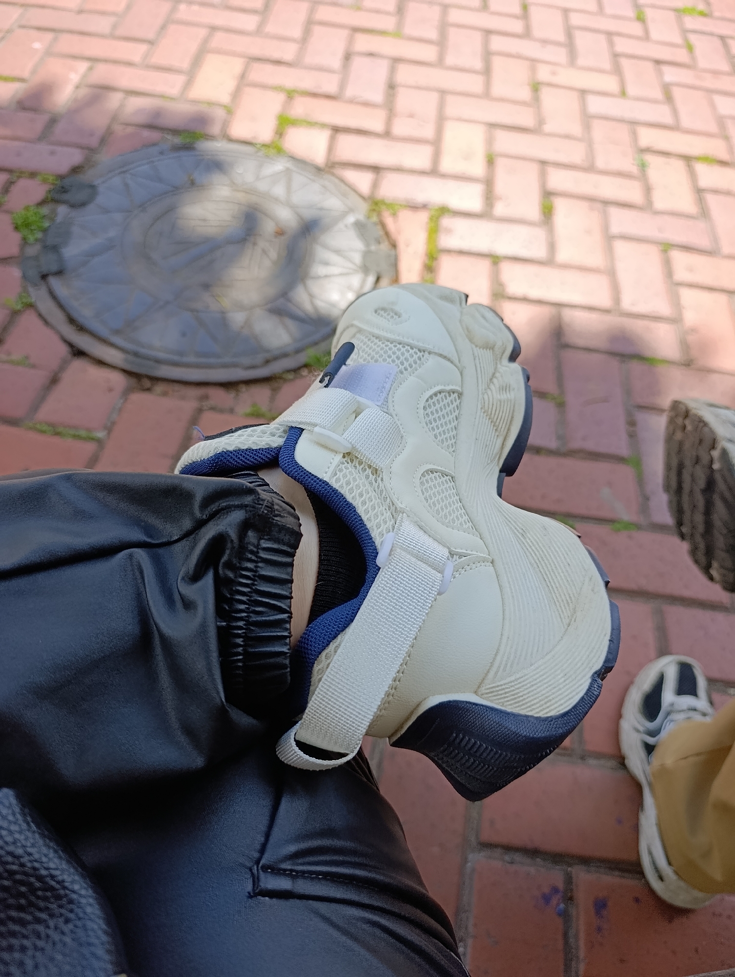The image shows a close-up view of a person's legs and feet, with a focus on a white and blue sneaker. The person is wearing black, shiny pants and a white sneaker with blue accents and white velcro straps. The sneaker is positioned on a brick-paved surface, and a circular metal manhole cover is visible in the background. Another person's feet, wearing a similar white and blue sneaker and light brown pants, are also visible in the lower right corner of the frame. The lighting suggests it is daytime, with shadows cast on the ground. The brickwork pattern on the ground is consistent with the type of paving commonly found on sidewalks and public spaces in Kyiv, Ukraine.